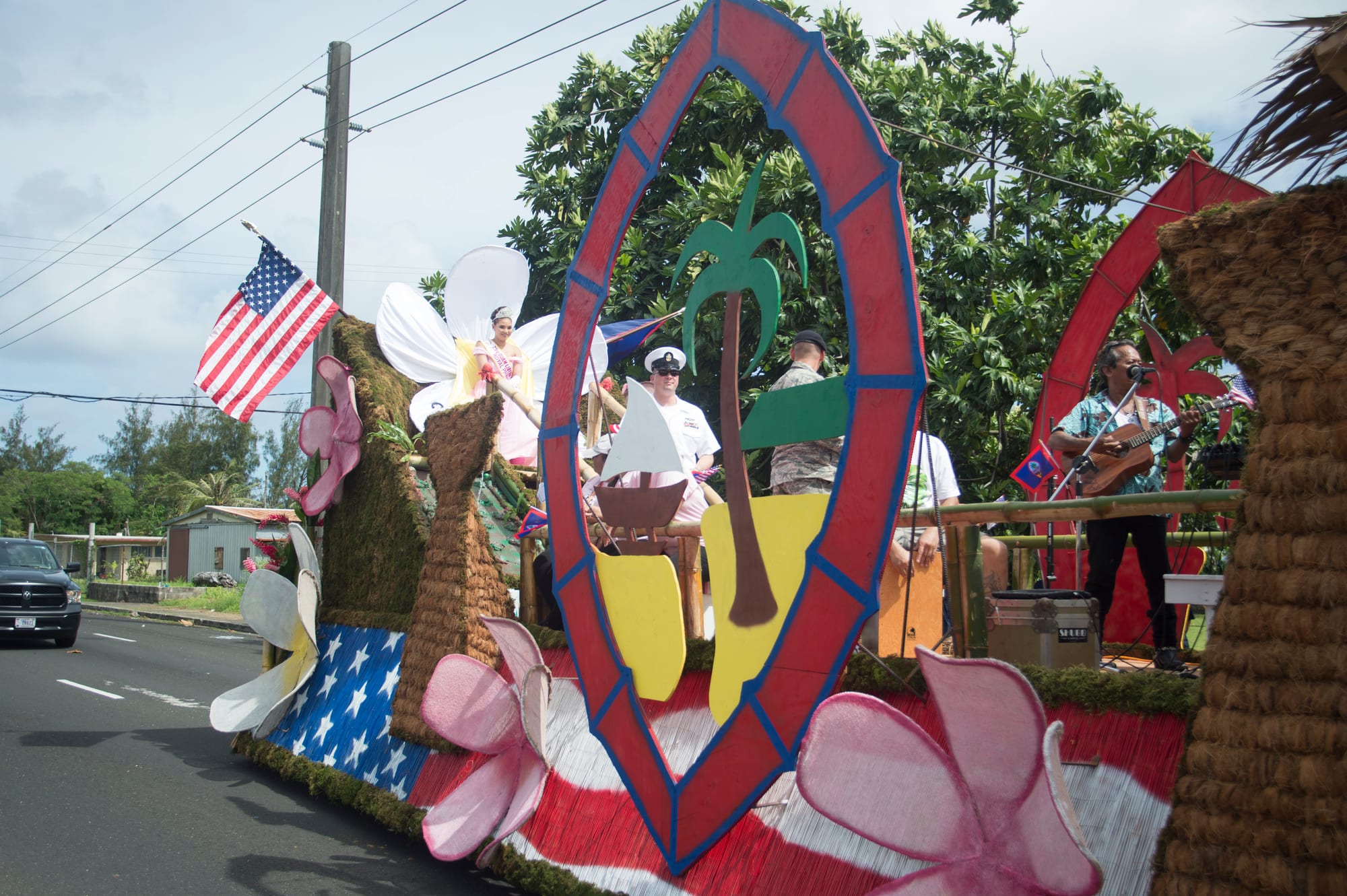 A parade float decorated with large pink and white flowers, a woven hut and a red oval arch framing a painted palm tree, carrying a woman in a white costume and a guitarist, with an American flag flying above
