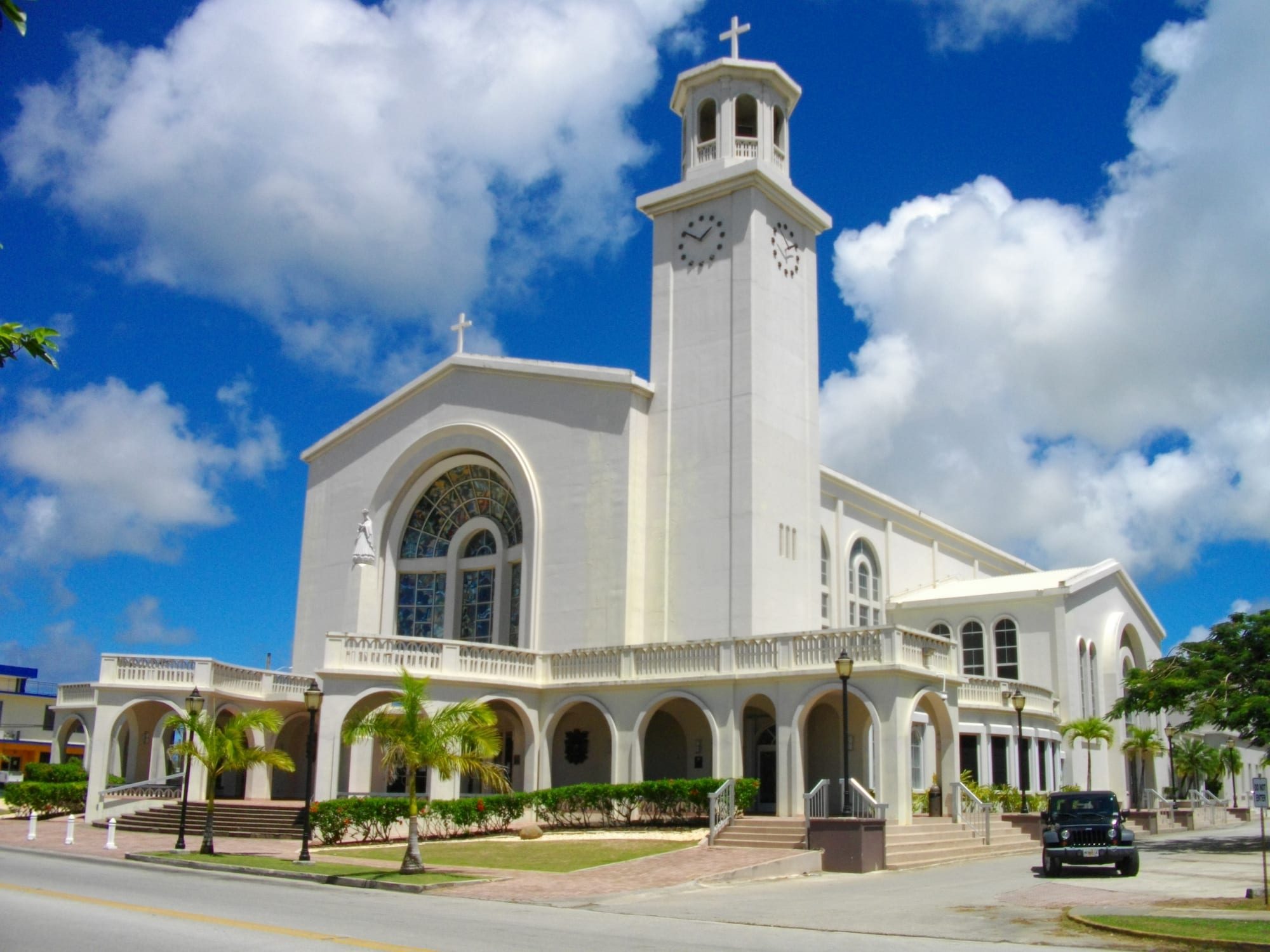 A large white church with a tall square clock tower topped by a cross, a big arched stained-glass window, and an arcaded balcony along the front, with palm trees and a lawn beneath a bright blue sky