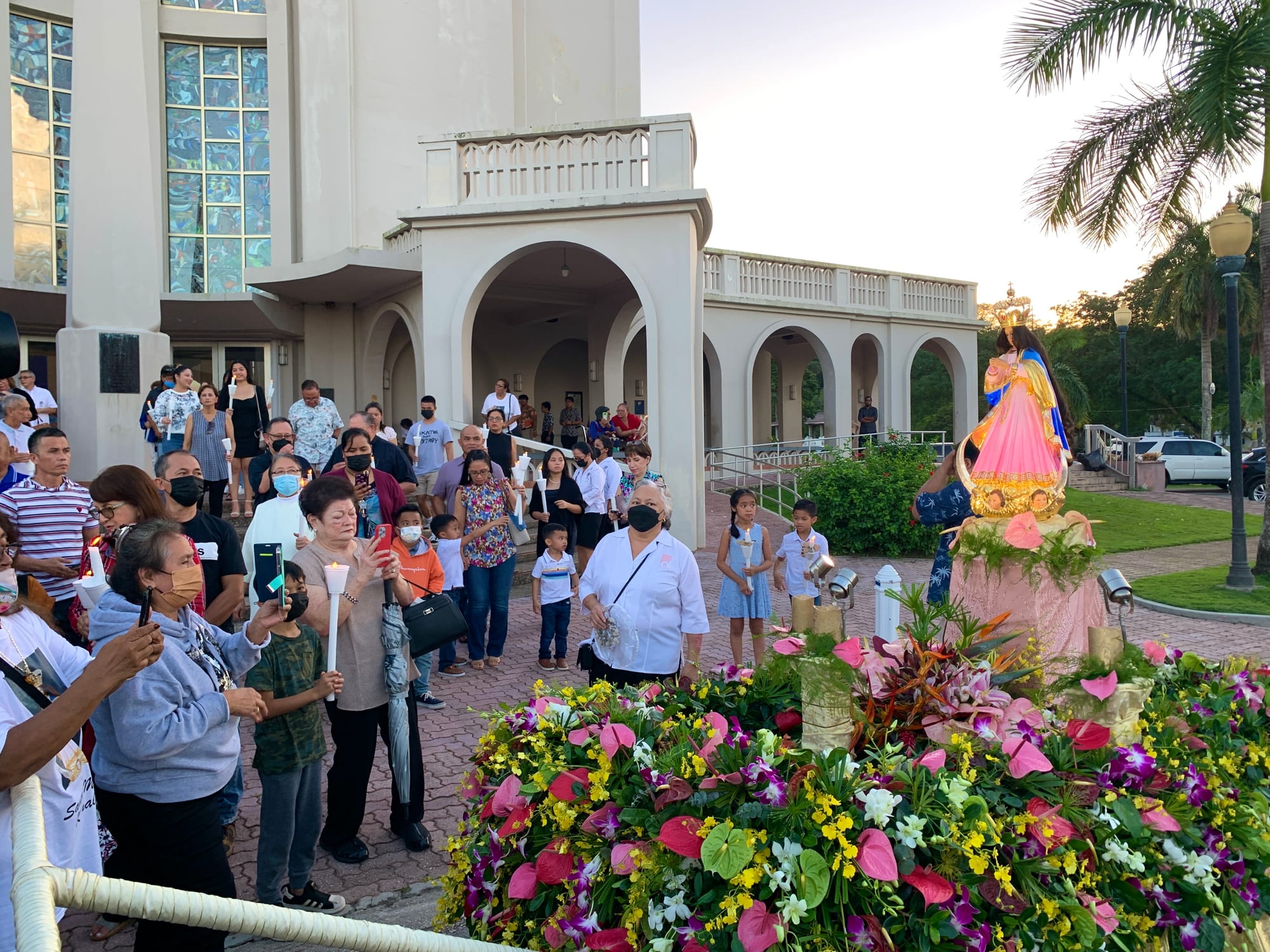 A crowd of people, some holding lit candles, gathers on a paved plaza beside a statue of the Virgin Mary in a pink and blue robe raised on a bed of bright tropical flowers, in front of a white church with arched arcades