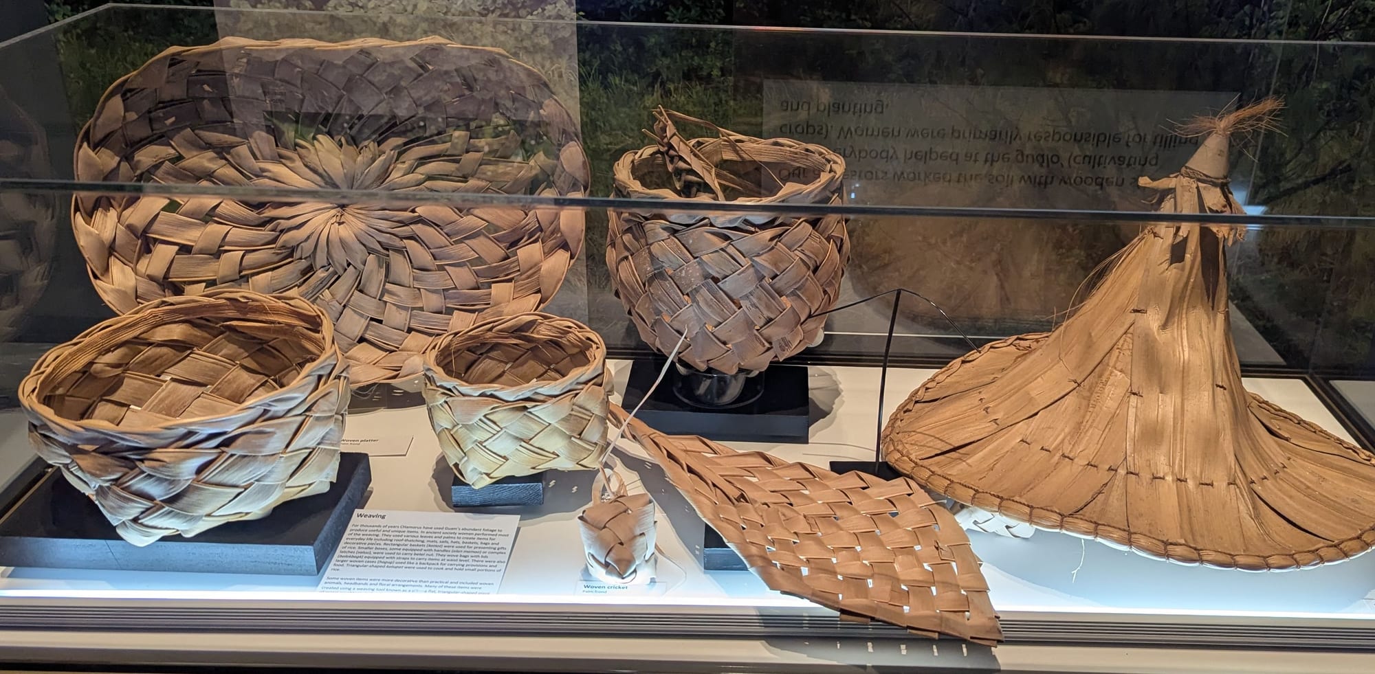 A museum display case holding several woven baskets of different sizes, a flat woven mat and a tall conical woven hat, all in pale natural leaf fibre