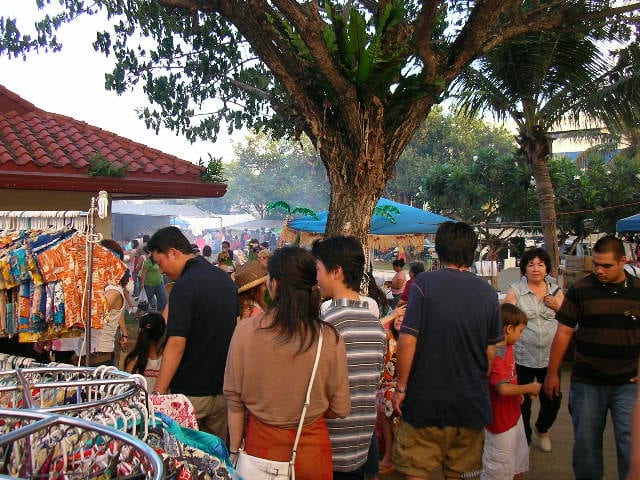 A crowd of shoppers browsing clothing racks and food stalls under palm trees at an outdoor evening market
