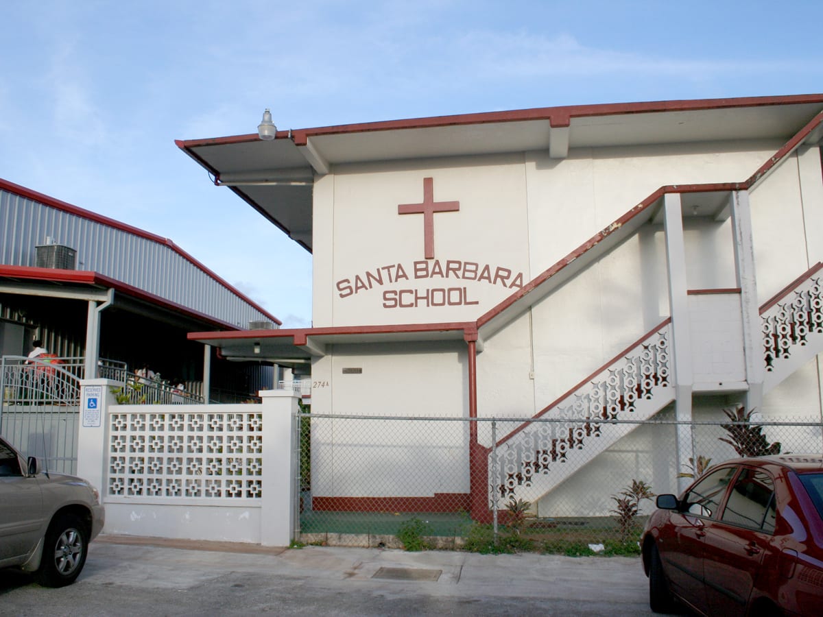 The white, red-trimmed Santa Barbara School building behind a chain-link fence, with two cars parked outside