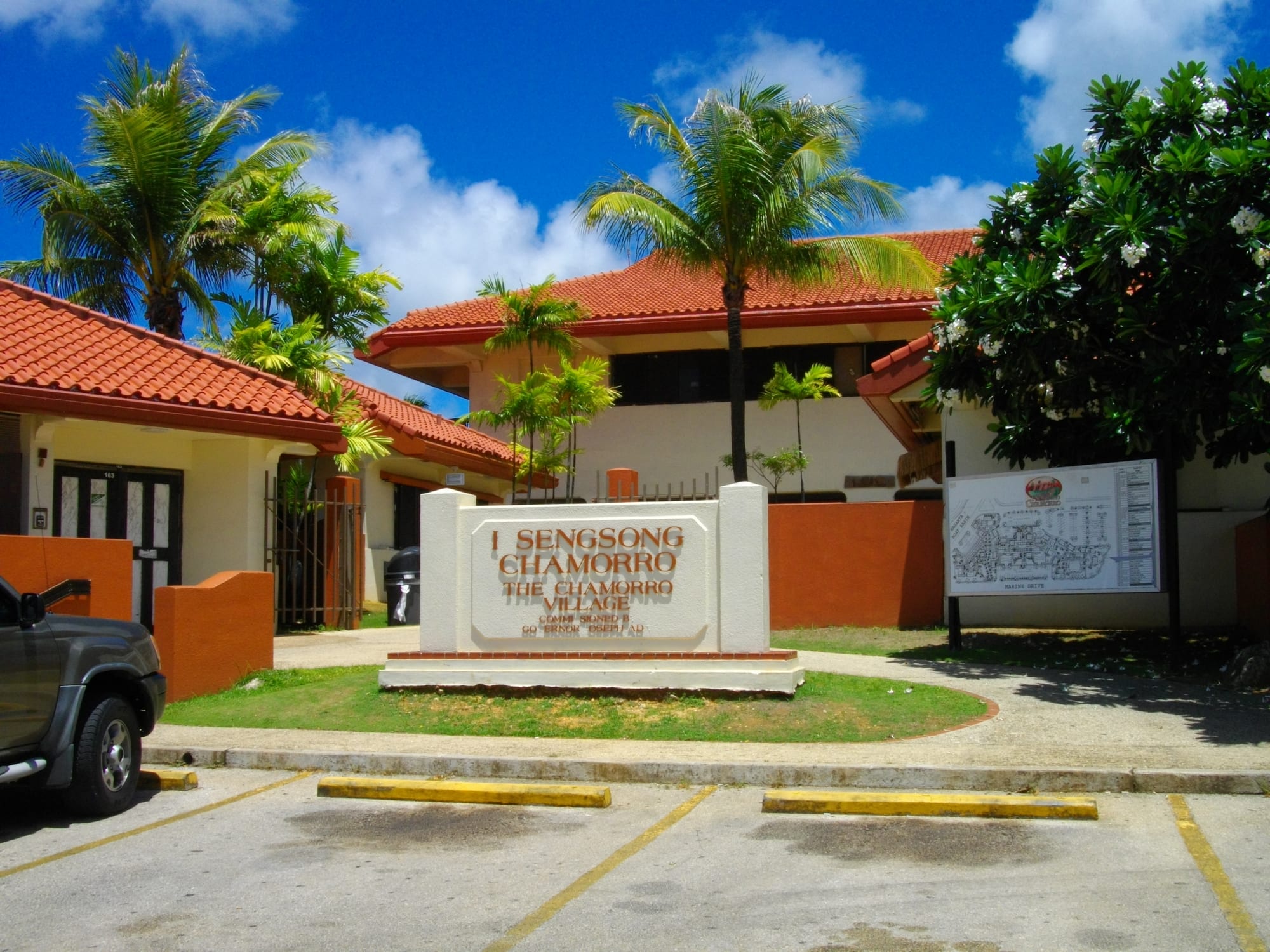 A tree-lined street beside a sign reading Sengsong Chamorro, The Chamorro Village, with red-roofed buildings behind