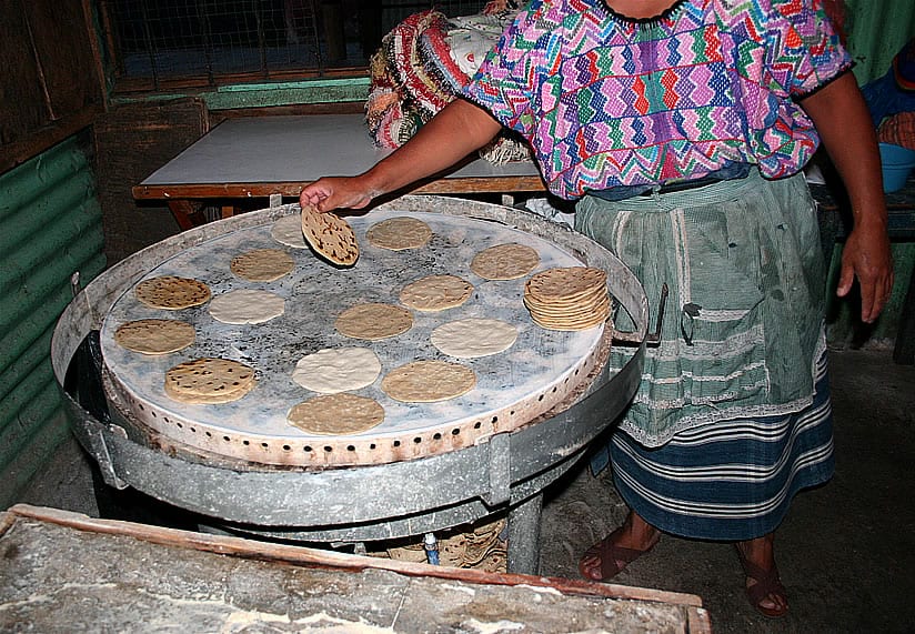 A woman in a traditional woven huipil and skirt cooking tortillas on a large round metal comal