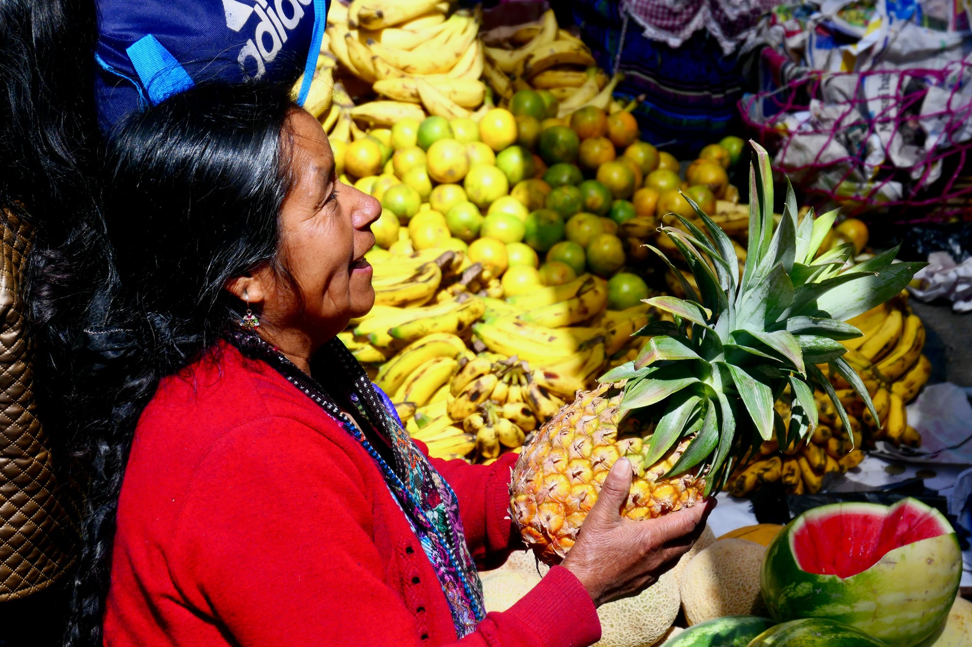 A woman holding a pineapple at a fruit stall piled high with bananas and citrus