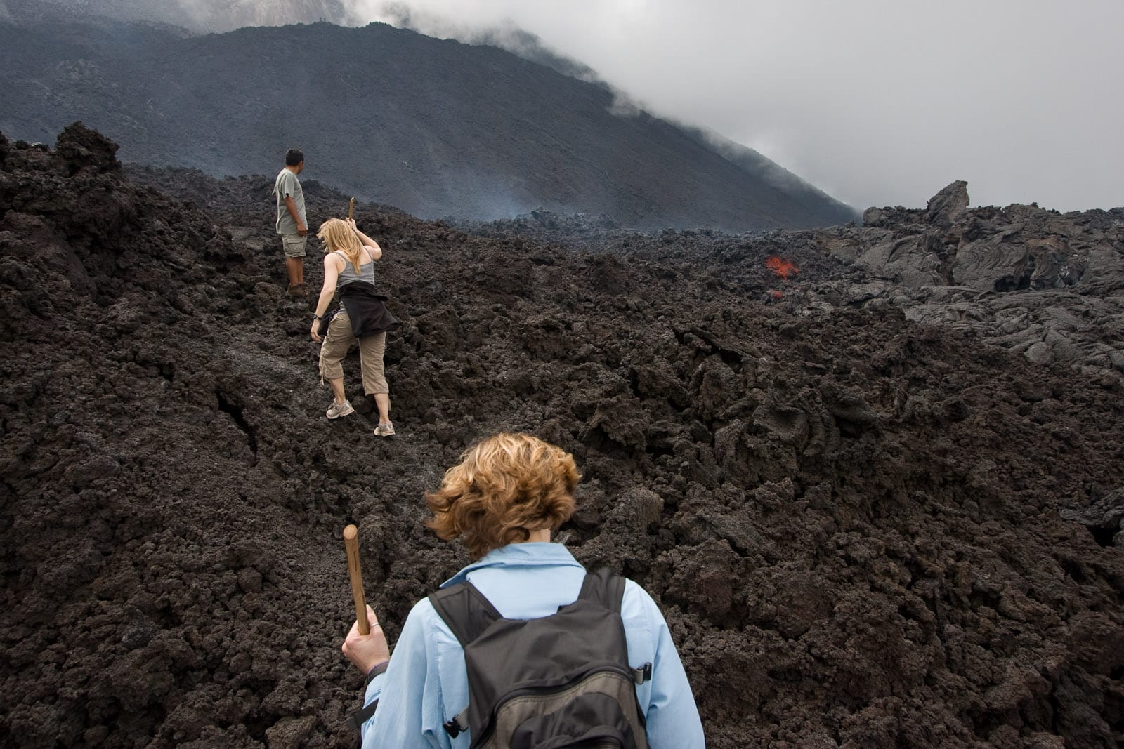Hikers crossing a black cinder field with glowing lava and steam ahead