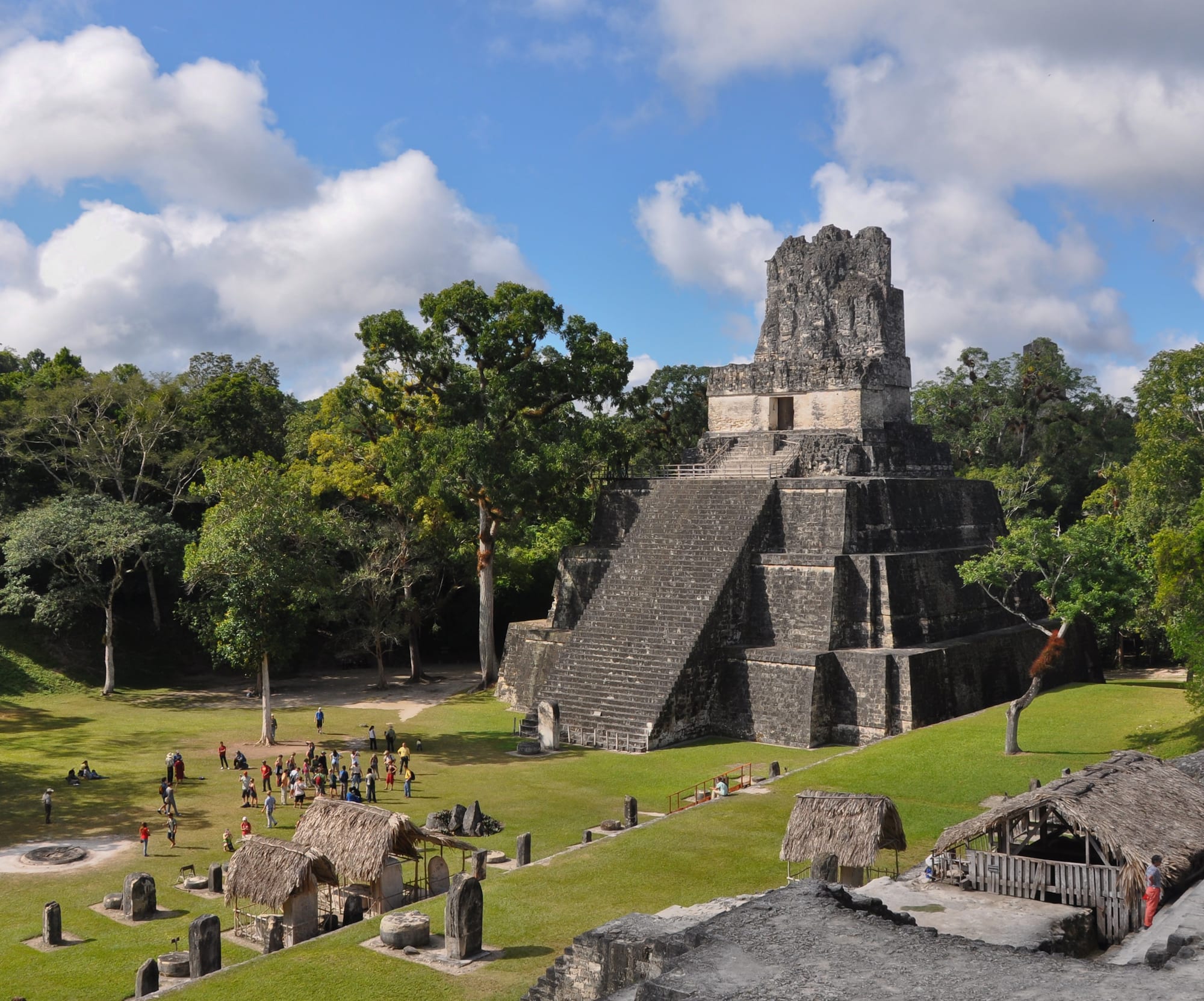 A steep grey limestone Maya pyramid with a temple on top, rising from cleared plaza and jungle