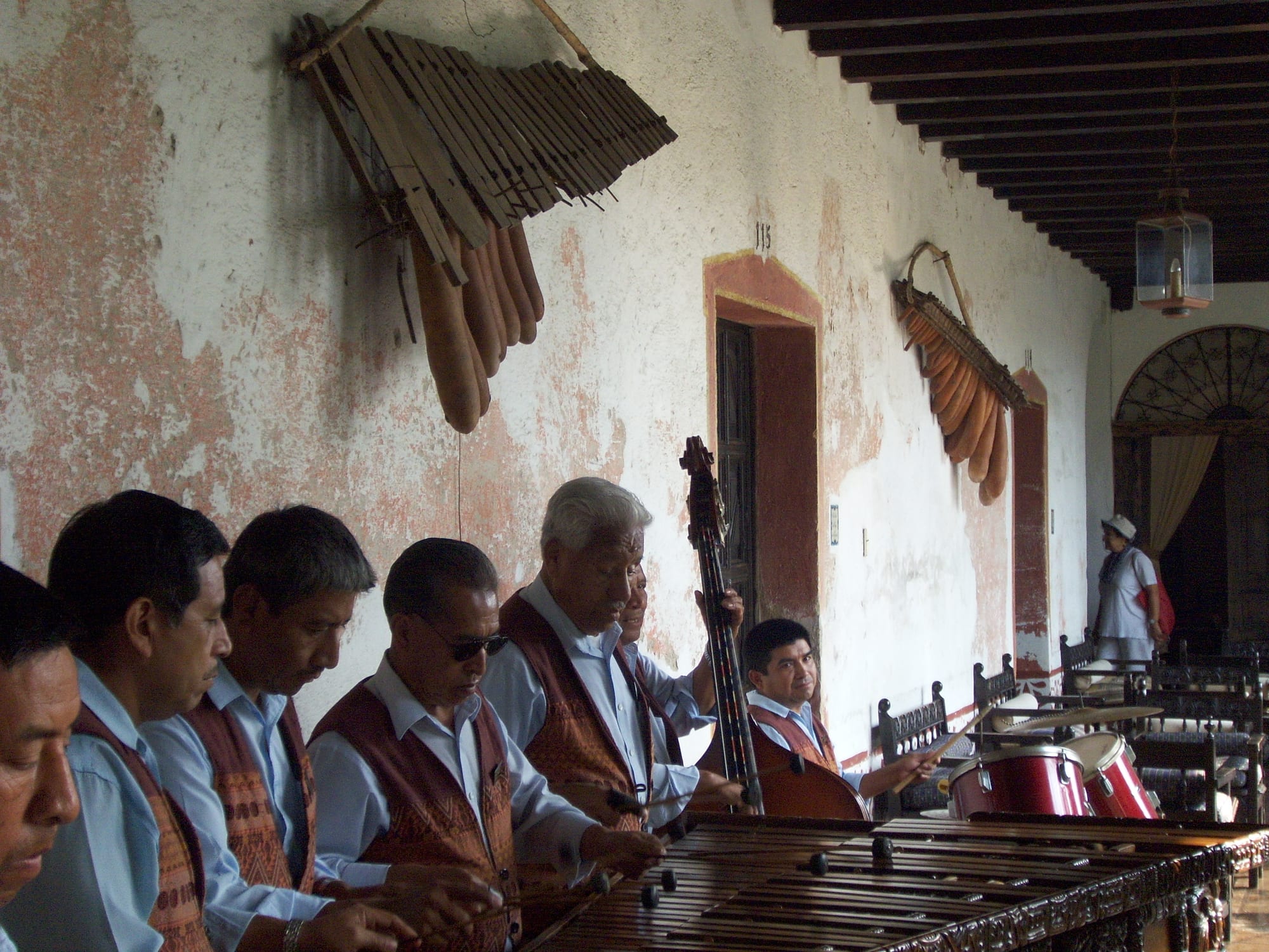 A row of men in pale blue shirts and patterned brown waistcoats play a long wooden marimba with mallets in a colonnaded courtyard, with old gourd-resonator marimbas hanging on the wall behind