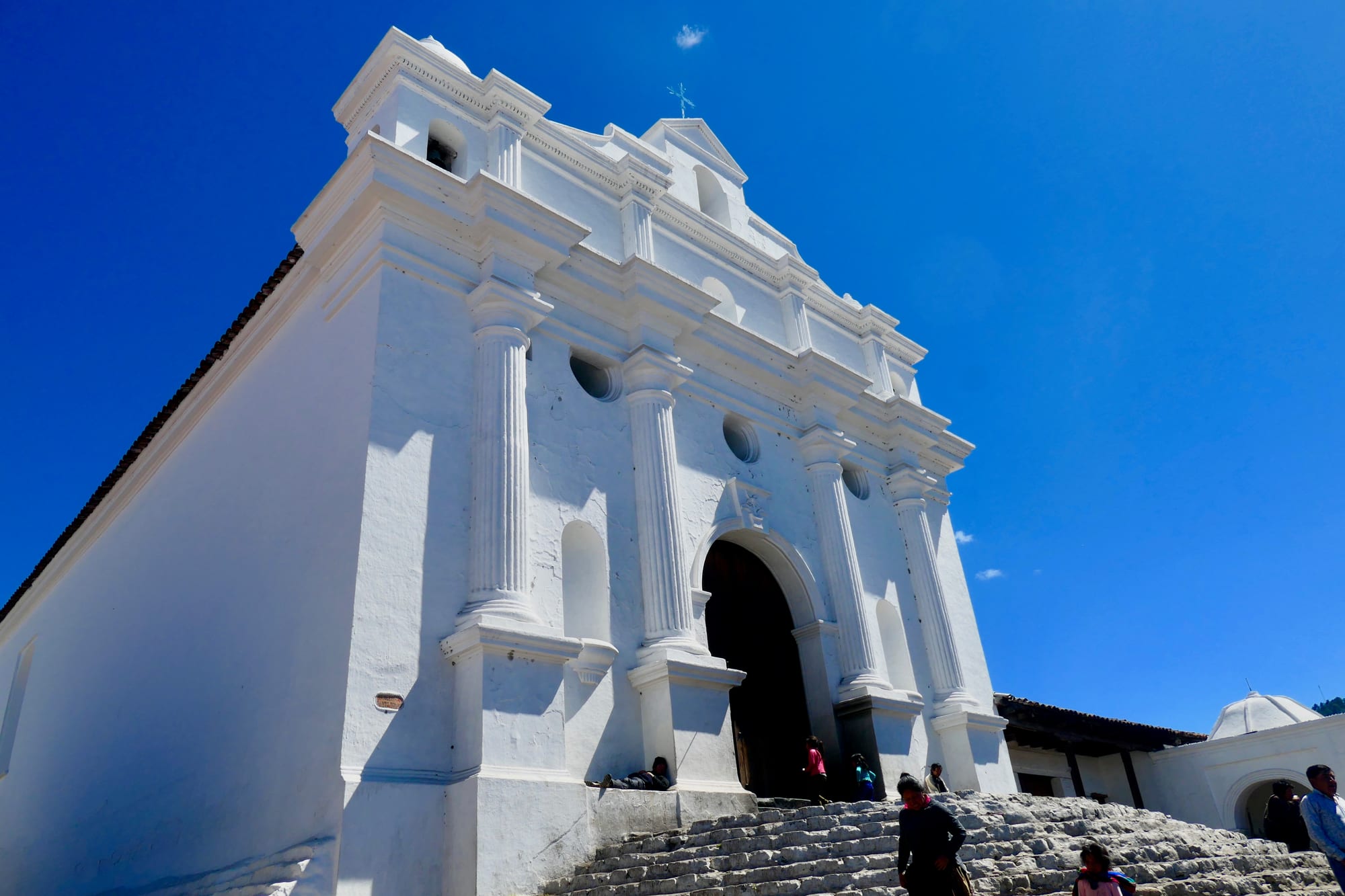 A whitewashed colonial church with fluted columns, round windows and a large dark wooden door, reached by a broad flight of worn stone steps, under a deep blue sky