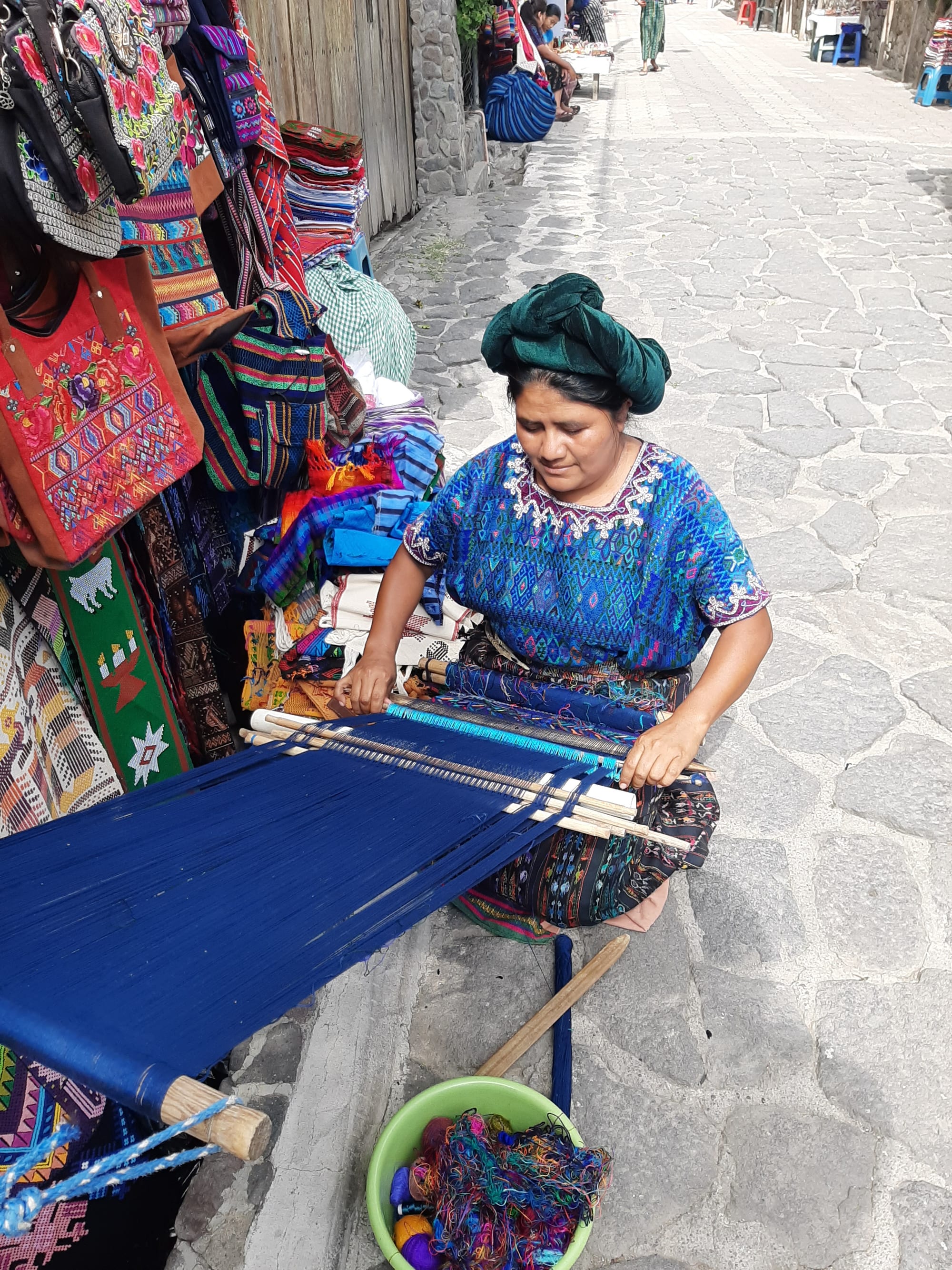 A woman in a bright blue embroidered blouse and a green headwrap sits on a stone street weaving deep blue thread on a loom stretched out in front of her, beside a stall hung with colourful bags and textiles