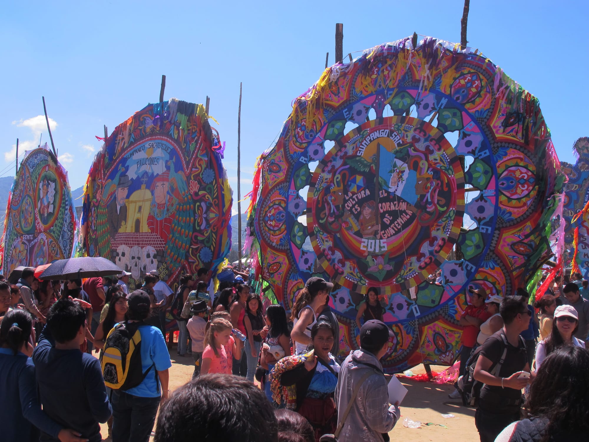 Several enormous round kites made of brightly coloured tissue paper stand upright on wooden poles above a crowd of people under a clear blue sky