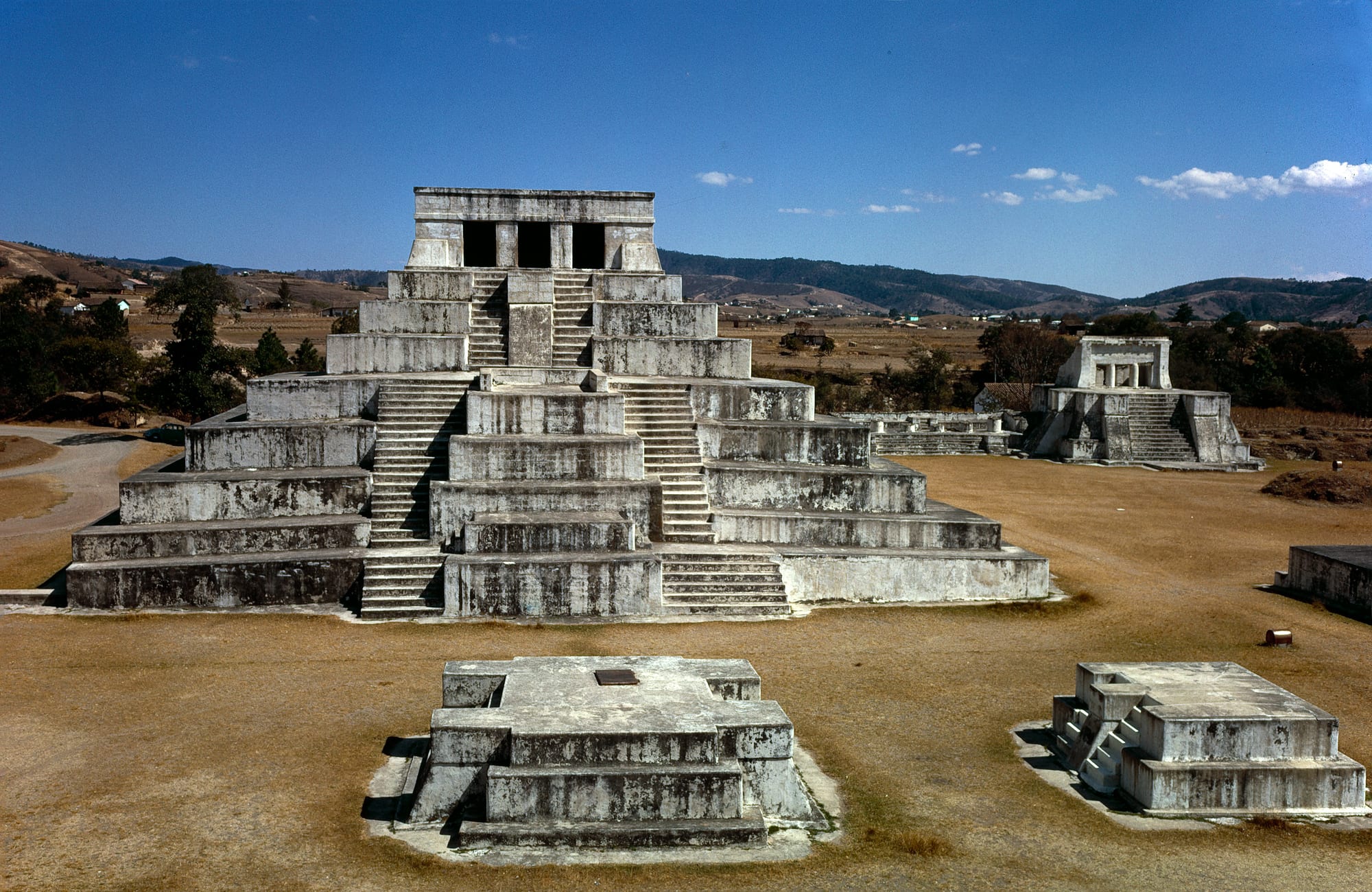 A white stepped pyramid with twin stairways and a columned shrine on top, standing in a dry grassy plaza with smaller platforms around it and hills behind