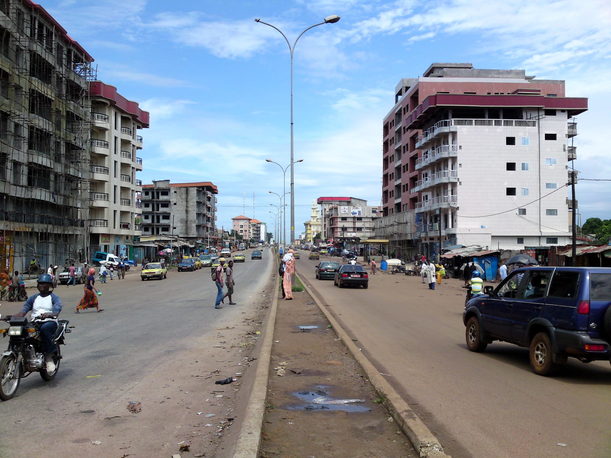 A busy, dusty city street with worn buildings, market stalls, and heavy traffic