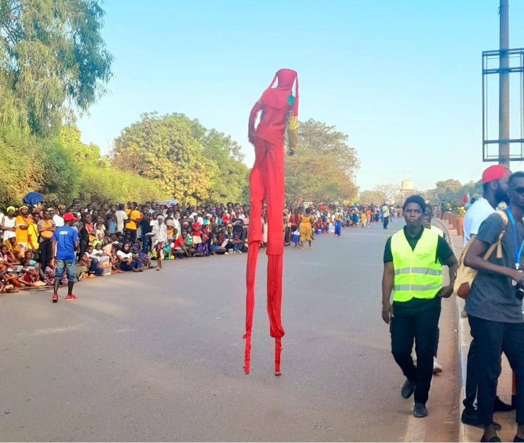 A performer on tall red stilts towers over a street crowd during a carnival parade