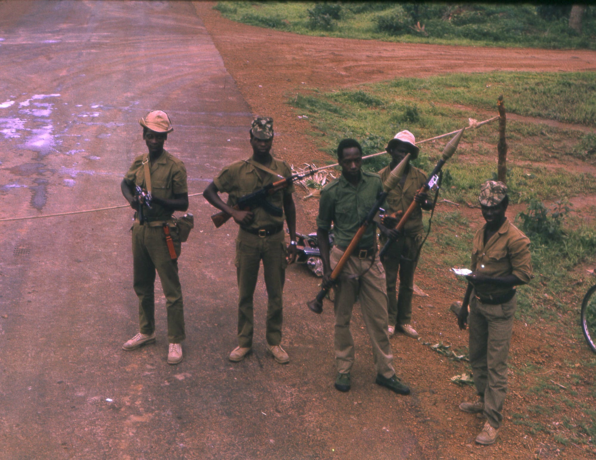 Five soldiers in olive uniforms and caps standing with rifles and rocket launchers beside a red-earth road, a rope stretched across it as a barrier