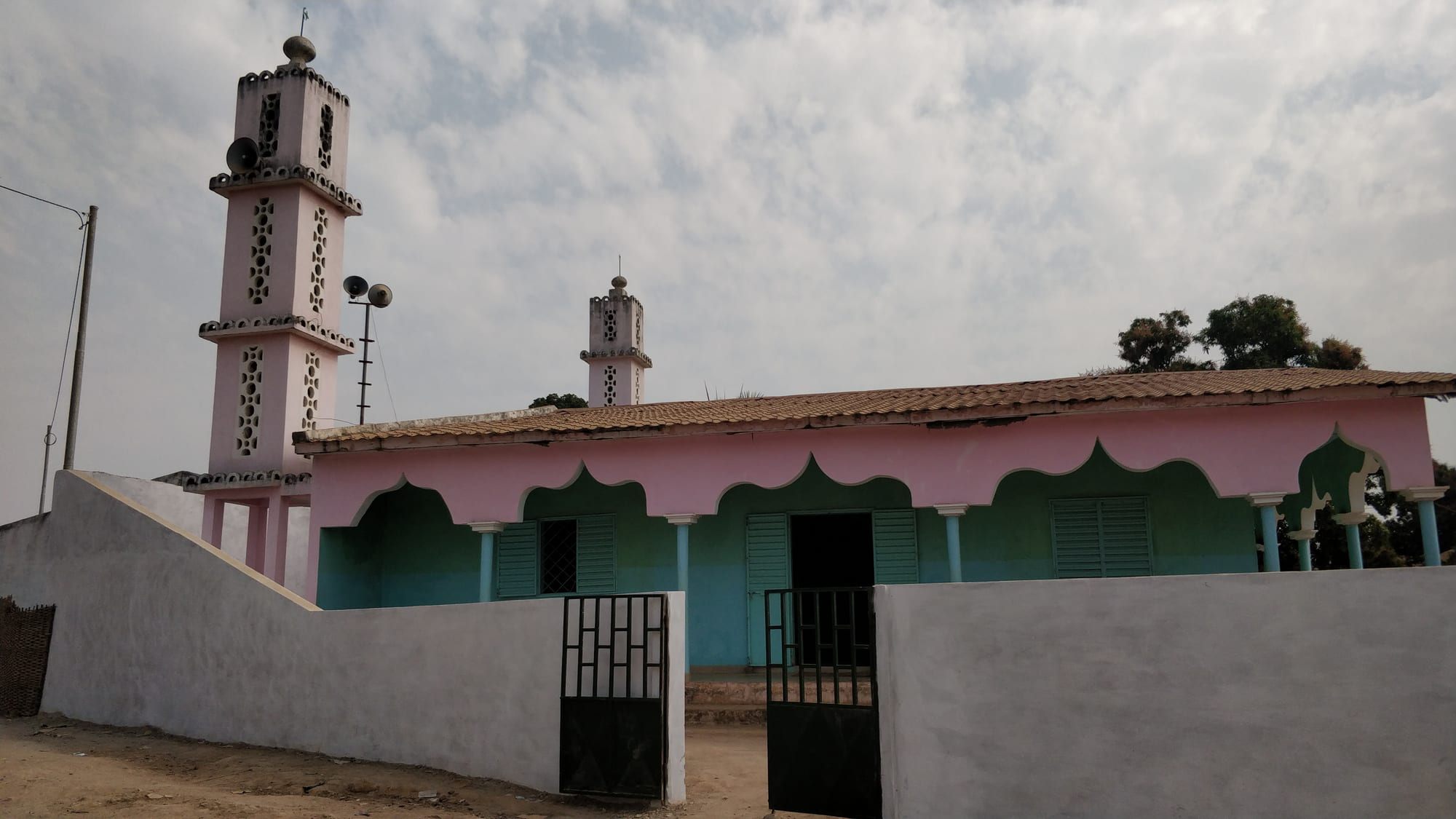 A low pink and green mosque with scalloped arches and two tall pink minarets fitted with loudspeakers, behind a white wall with black metal gates