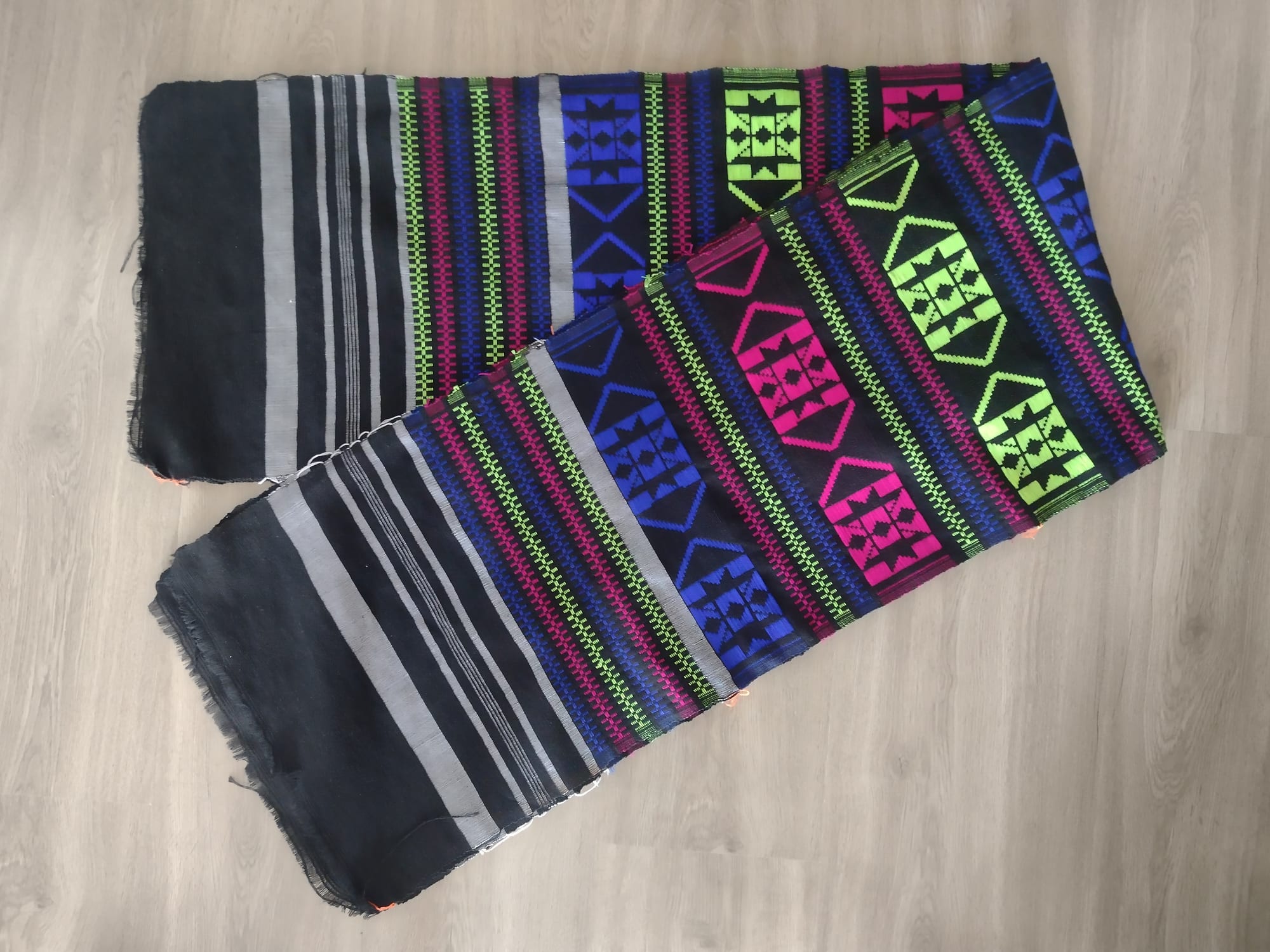 A folded length of handwoven cloth in black and grey stripes with bands of bright blue, pink and lime-green geometric patterns, lying on a wooden floor
