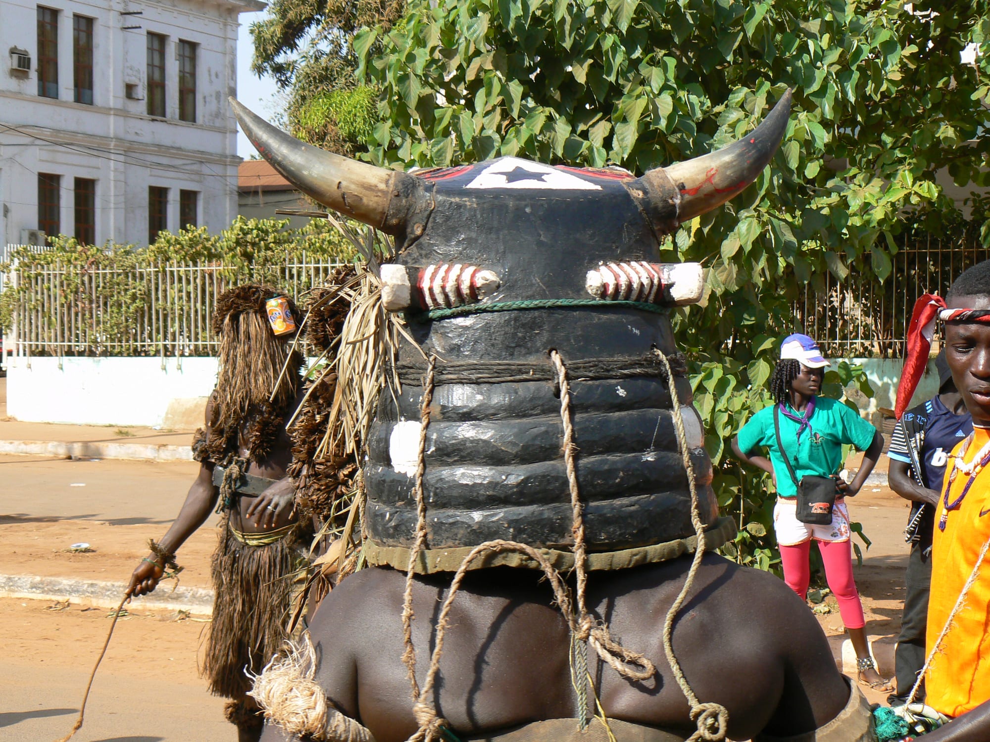 A performer seen from behind wearing a large black horned mask bound with rope, with a costumed figure in a fibre outfit and spectators on a street beyond