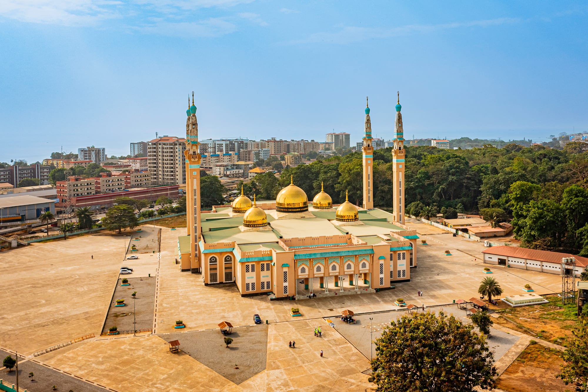 An aerial view of a large golden-domed mosque with four tall minarets, set among trees with the city and sea beyond