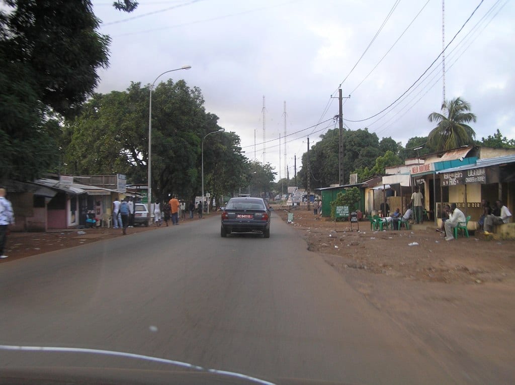 A residential Conakry street with roadside shops, parked chairs, and people sitting and talking outside