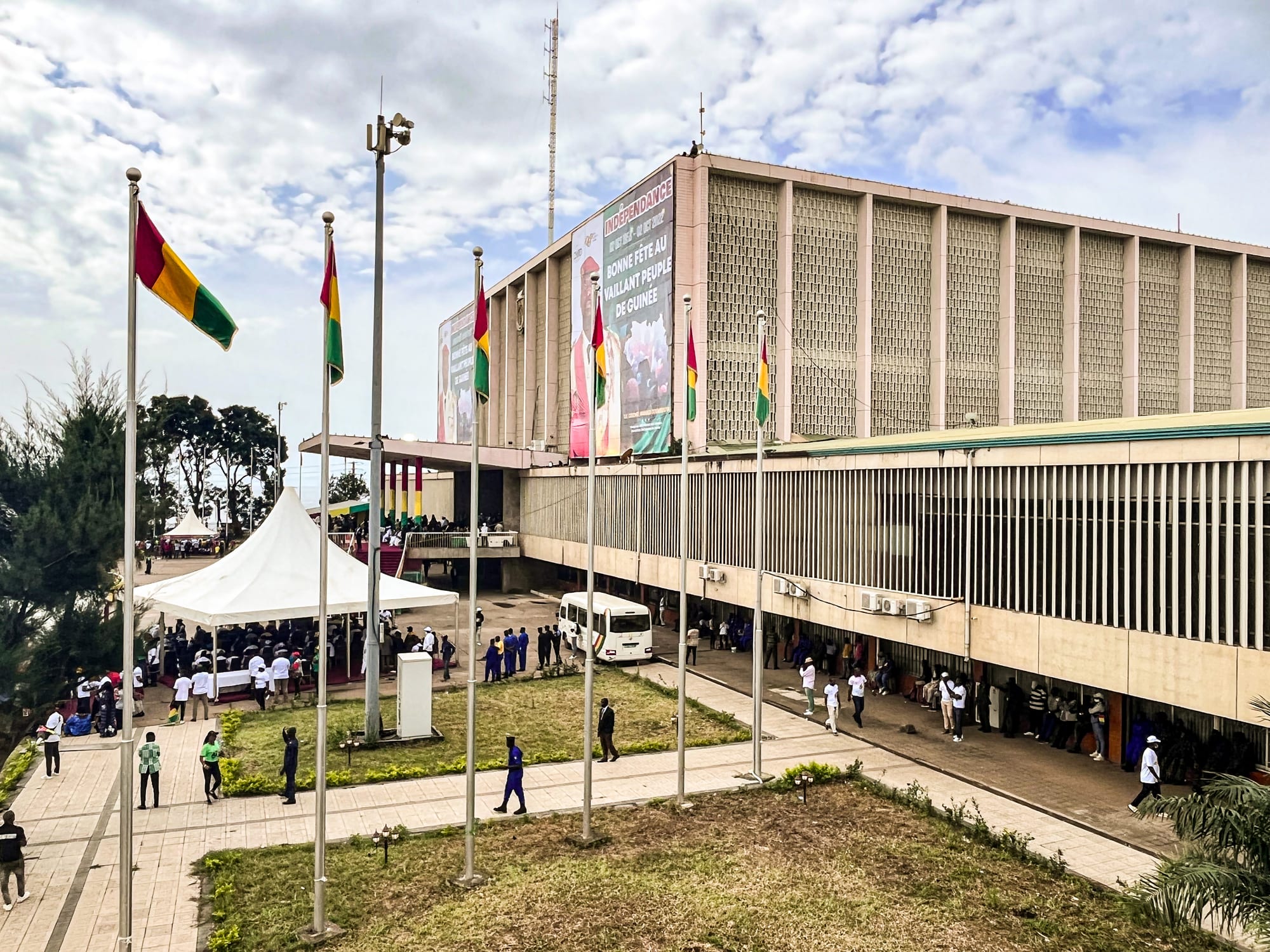 Red, yellow and green Guinean flags on tall poles in front of a large concrete public building hung with an independence-day banner, with a crowd gathered under a white tent