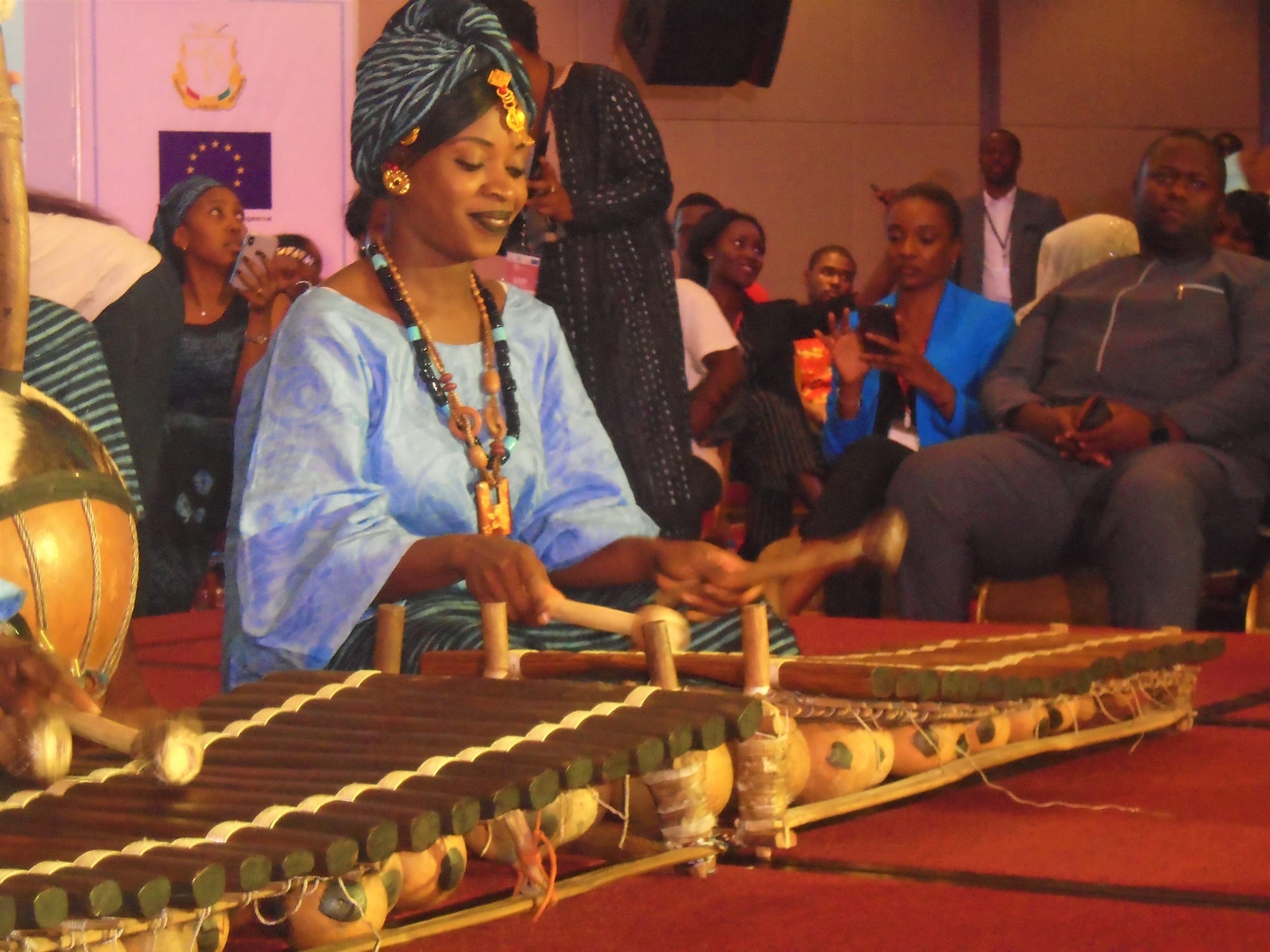 A woman in a pale blue robe, patterned headwrap and bead necklaces smiles as she strikes the wooden keys of a balafon, with gourd resonators hanging beneath and an audience seated behind her