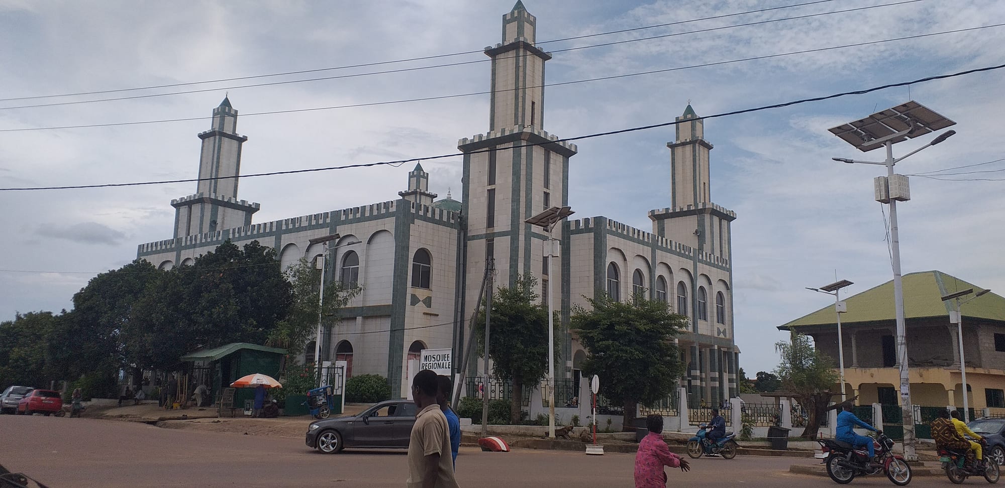 A large white mosque with green trim, arched windows and four tall square minarets, beside a road with motorbikes, a parked car and people walking