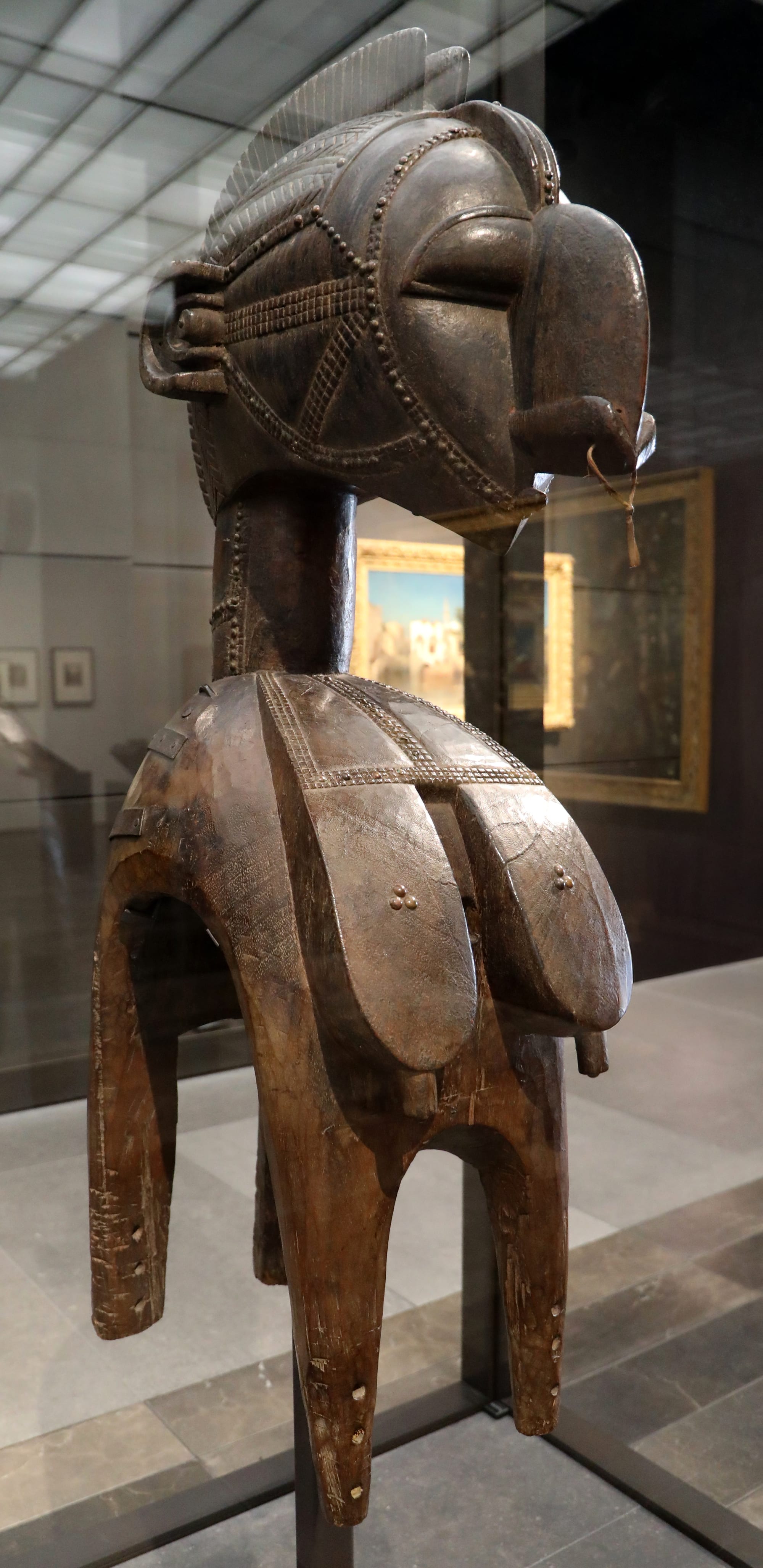 A tall dark wooden headdress in a museum case, with a large carved head and crested hair on a slender neck, above a bust and four curved legs