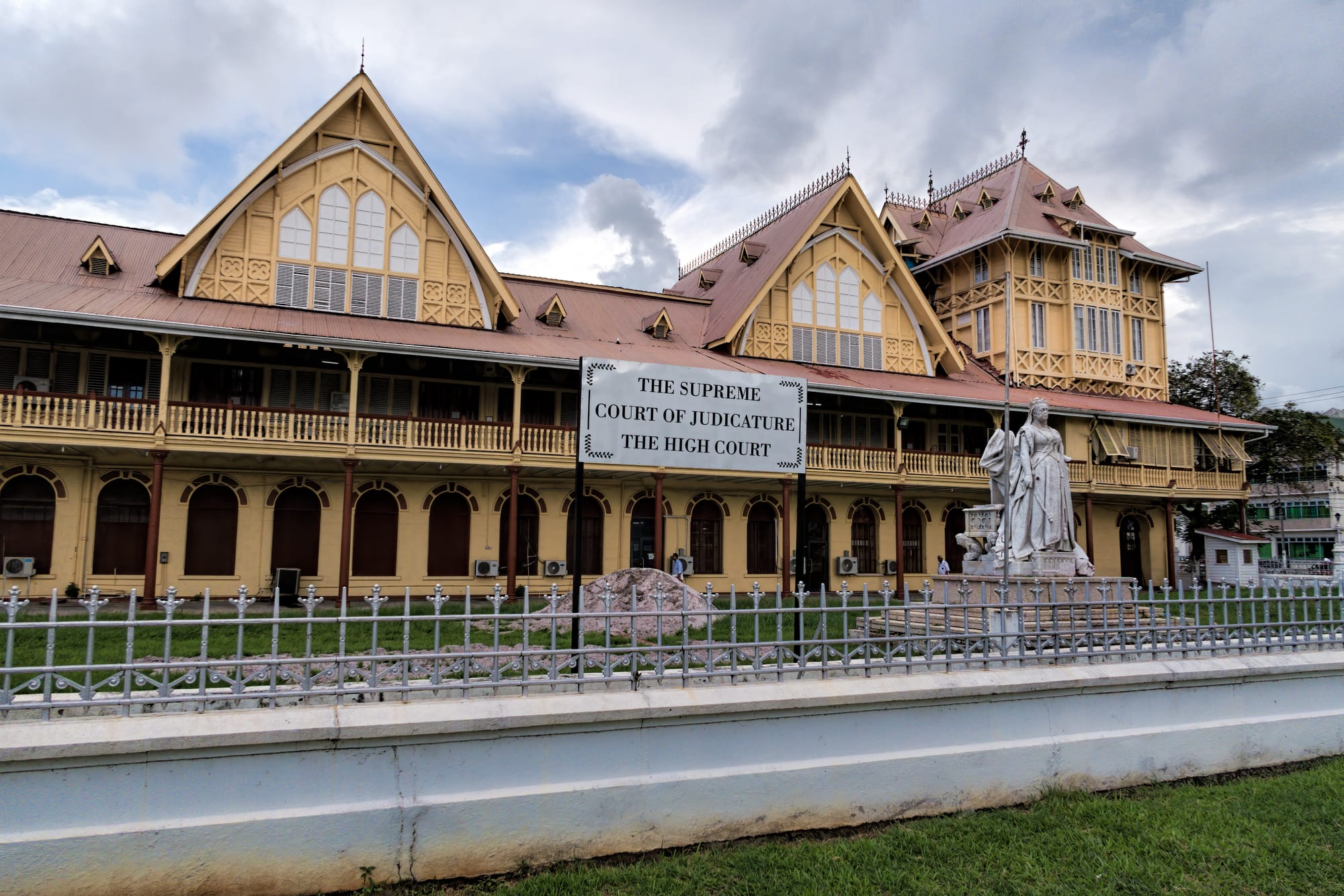 A long yellow timber-and-masonry court building with steep red gabled roofs, a sign reading The Supreme Court of Judicature, The High Court, and a white marble statue of Queen Victoria in front