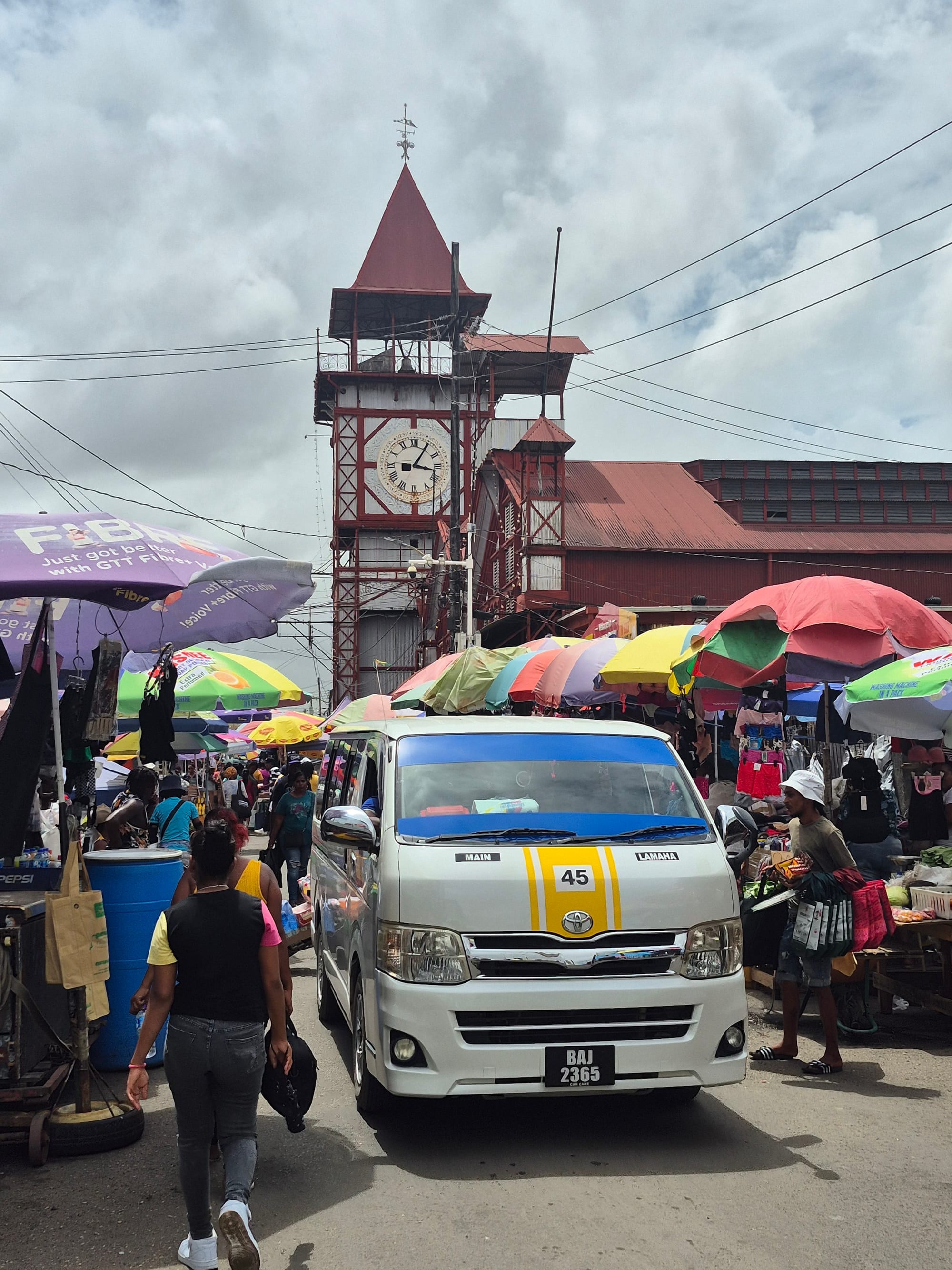 A minibus parked near a red clock tower and colourful market umbrellas at Stabroek Market