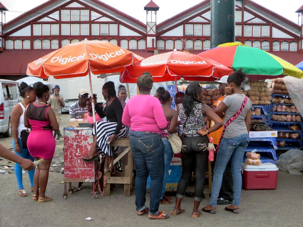 Women shopping at a street stall with umbrellas outside a large red-trimmed market building