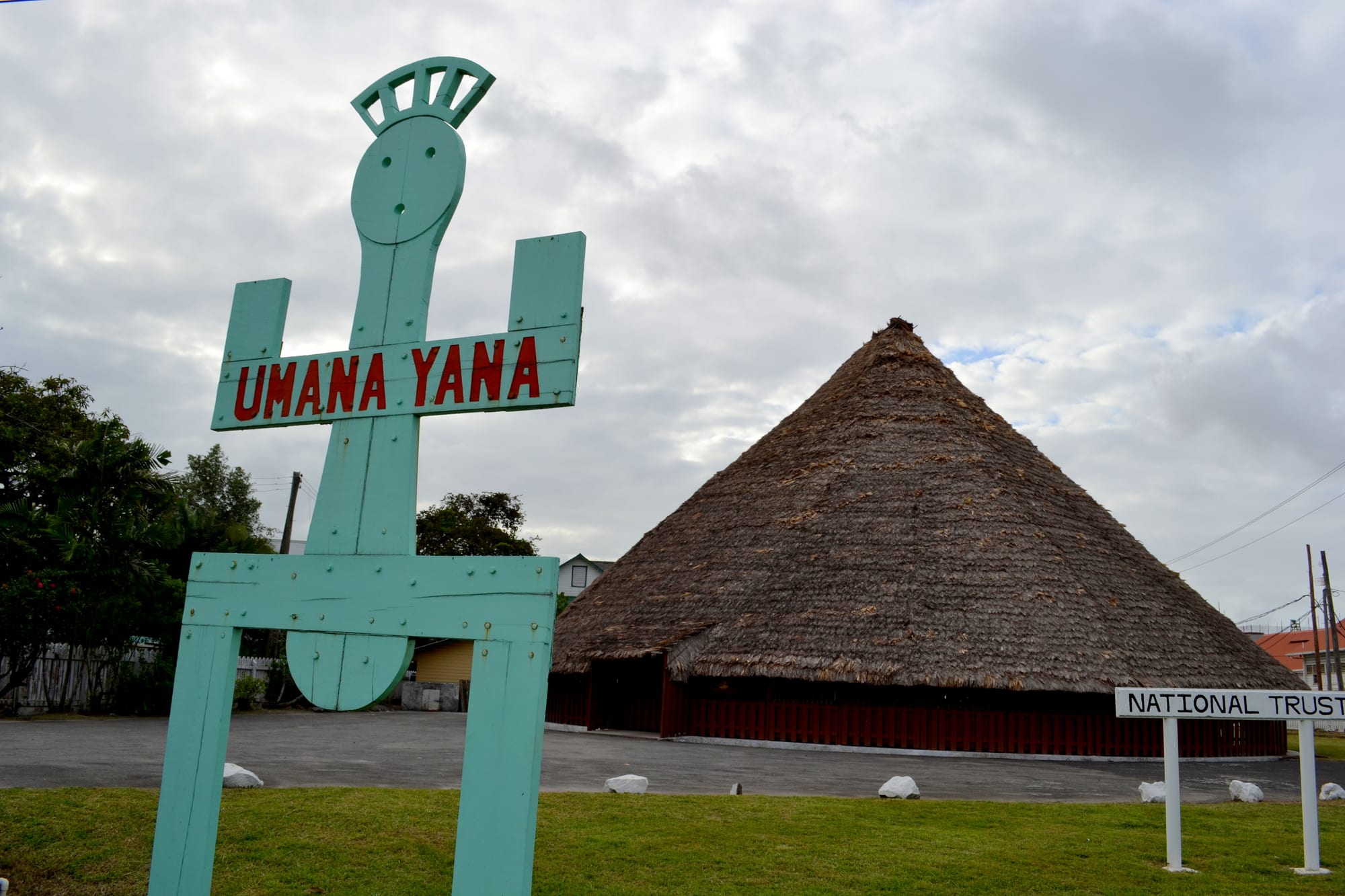 A tall conical building with a thick thatched roof over red wooden walls, beside a turquoise wooden sign spelling Umana Yana and a smaller National Trust sign on a lawn