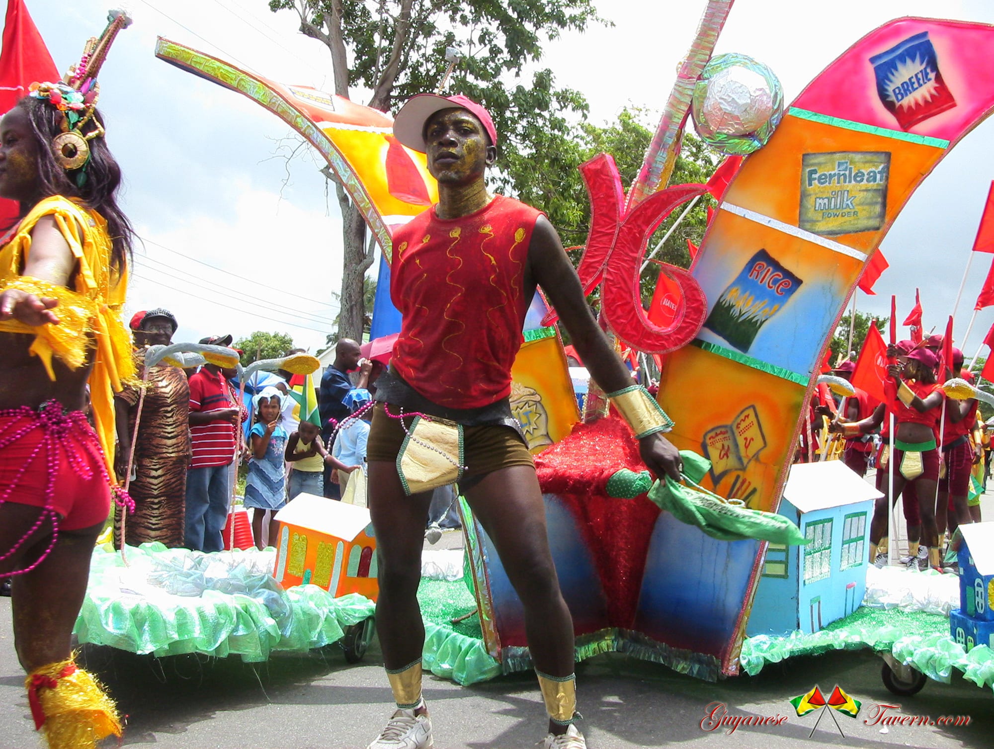 A costumed reveller in a red top and gold arm cuffs stands in front of a brightly painted float decorated with model houses and pictures of everyday goods, with other costumed performers and spectators around it