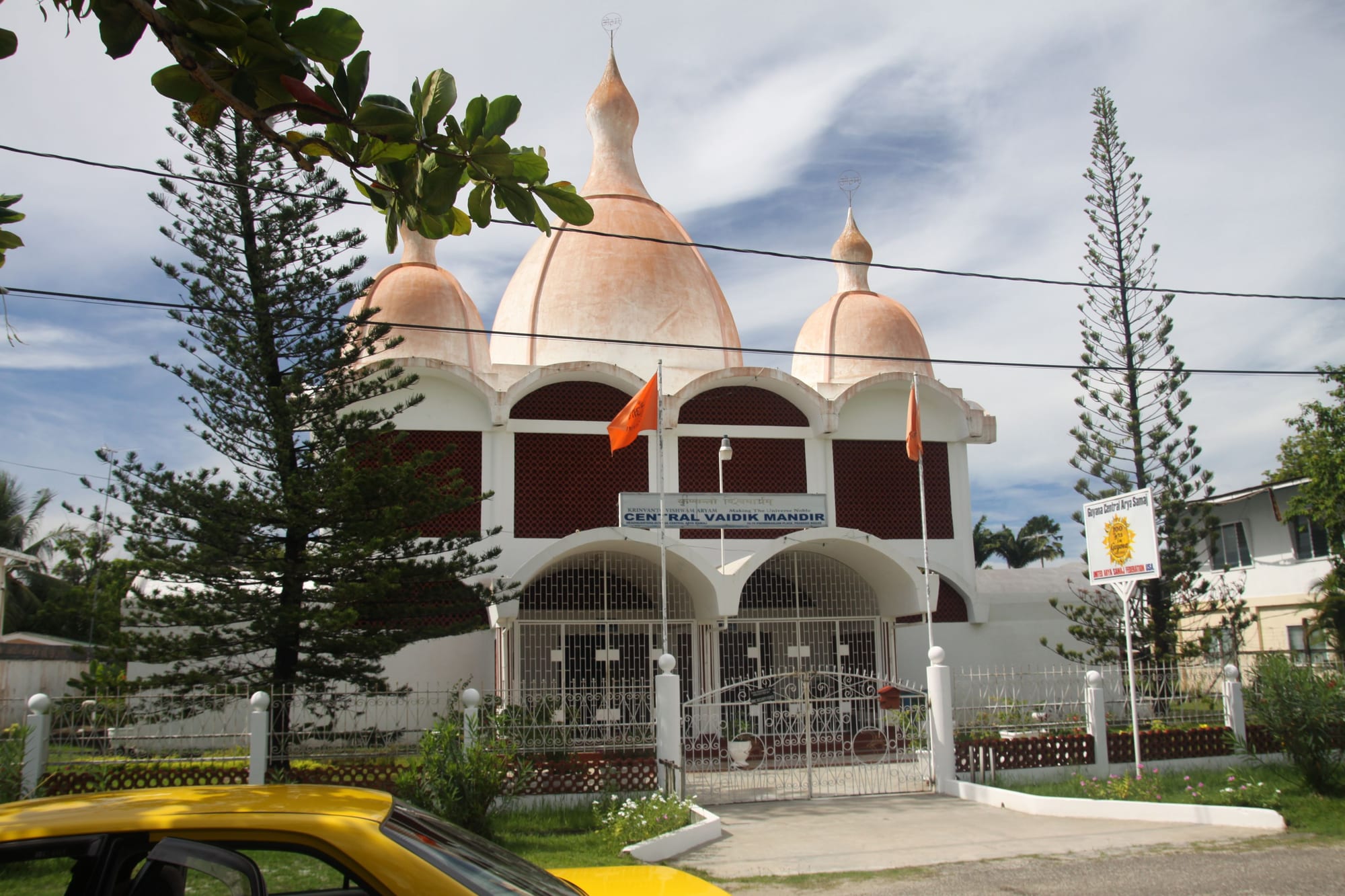 A white Hindu temple with three pale pink onion-shaped domes, arched windows and orange flags, behind a white fence with a gate and tall pine trees on either side