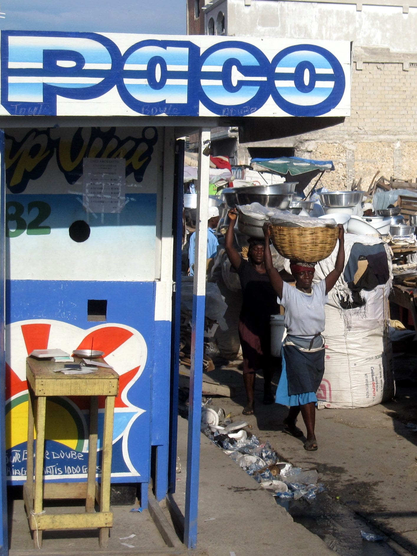 Two women carrying large basins and a basket balanced on their heads through a market alley