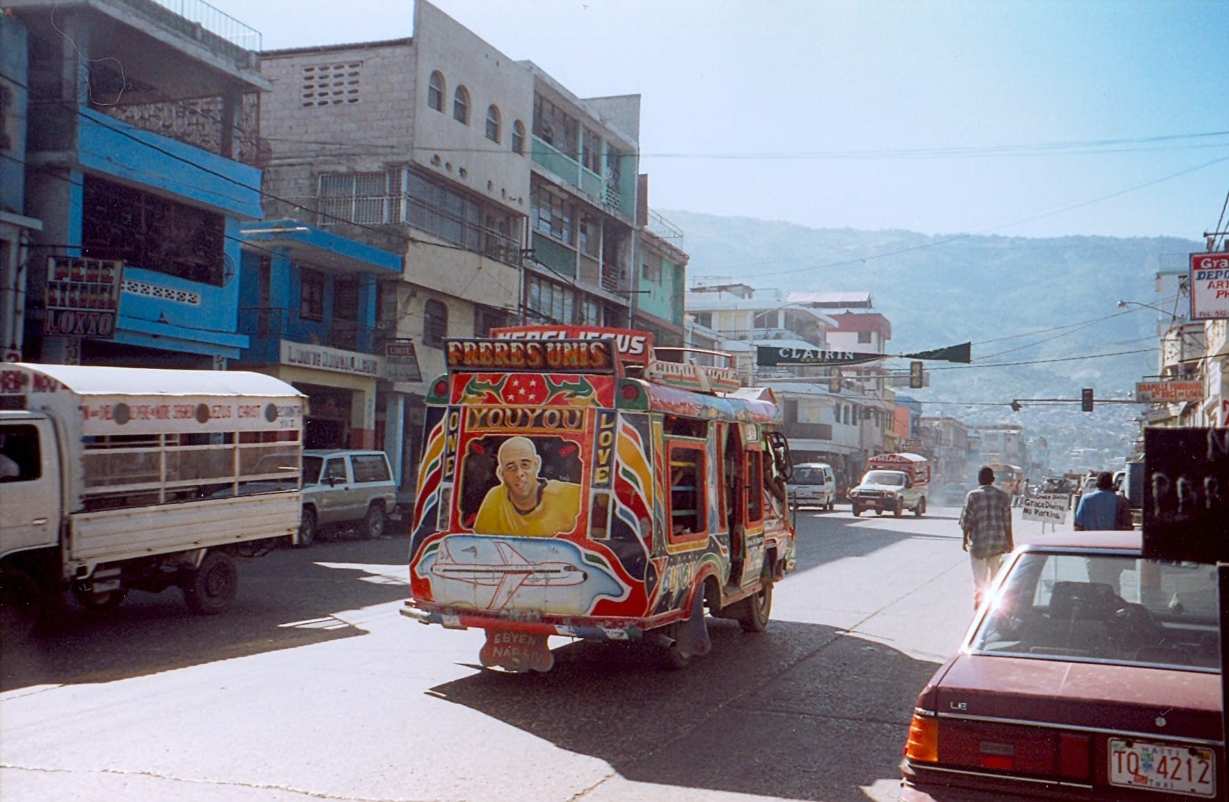 A brightly painted tap-tap truck with murals and lettering, driving down a street lined with colourful buildings