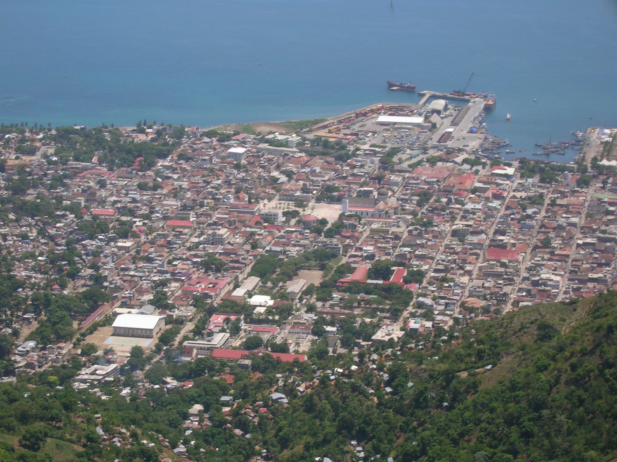 An aerial view of a coastal city's grid of streets and rooftops beside a harbour