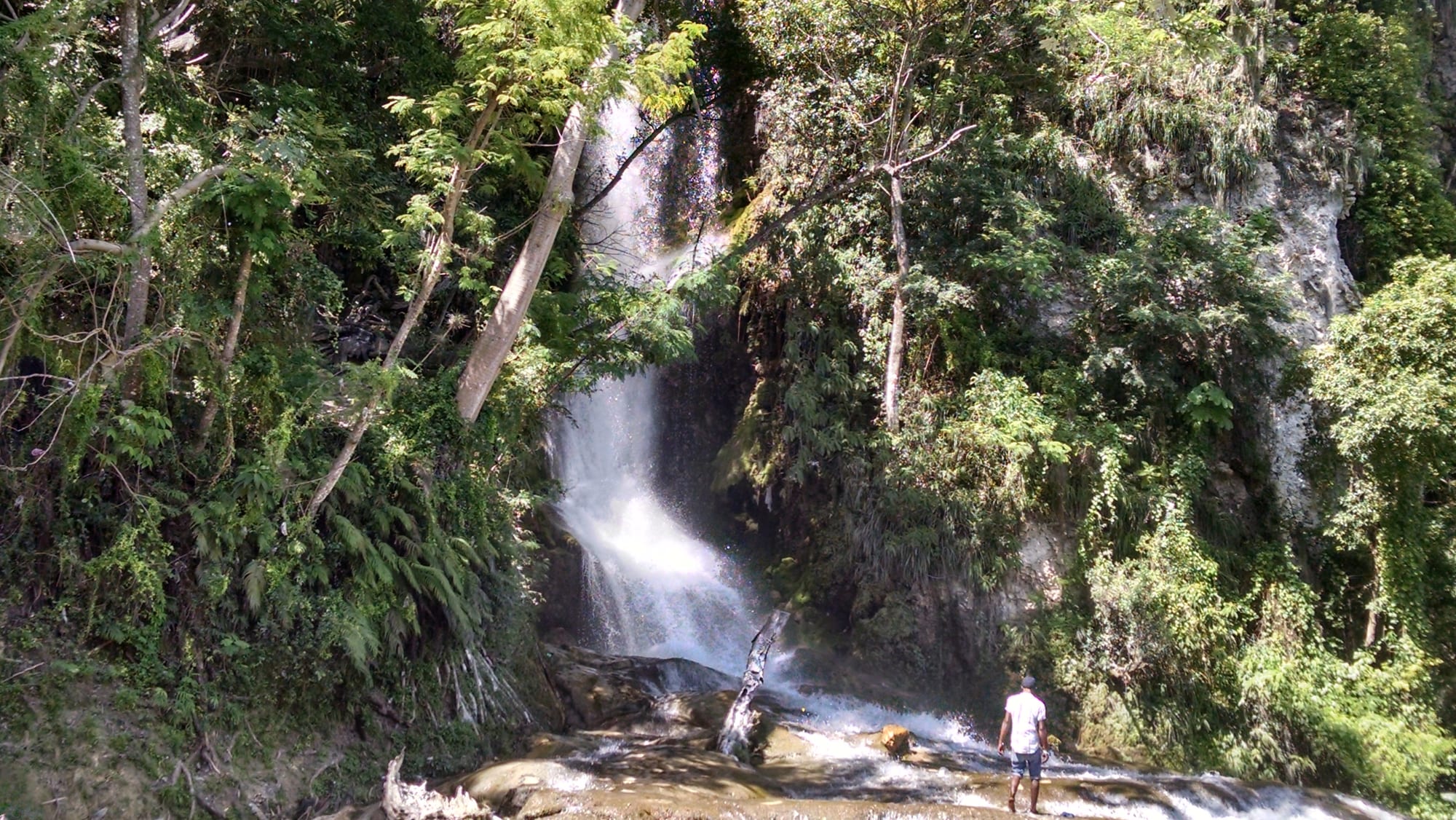 People bathing in a wide, powerful waterfall spilling over greenery-covered rock