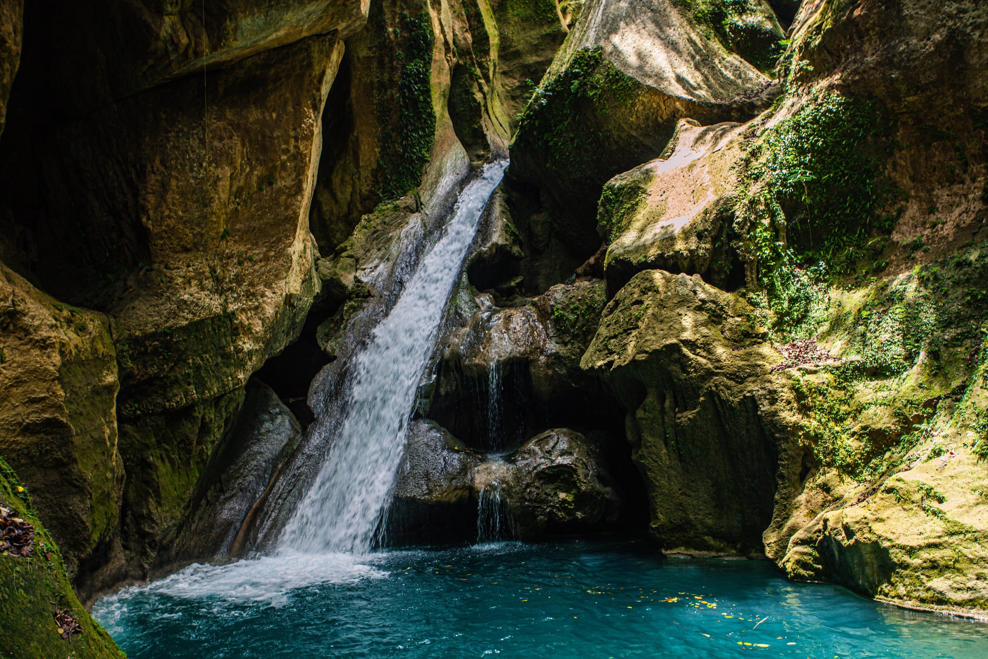 A deep blue-green pool at the base of a mossy rock cliff, fed by a small waterfall