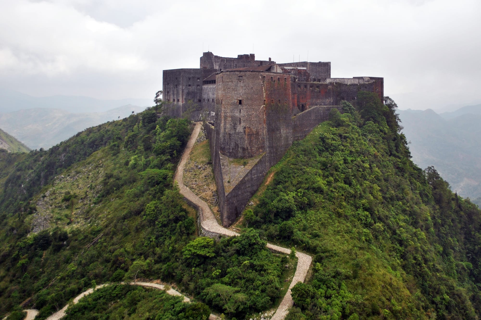 A massive stone fortress with pointed prow-like walls on top of an isolated green mountain