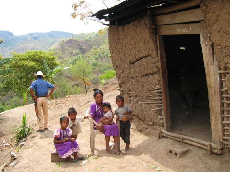 A mother and three children standing outside a mud-brick house with a corrugated tin roof in rural Honduras, with a man and hillside in the background