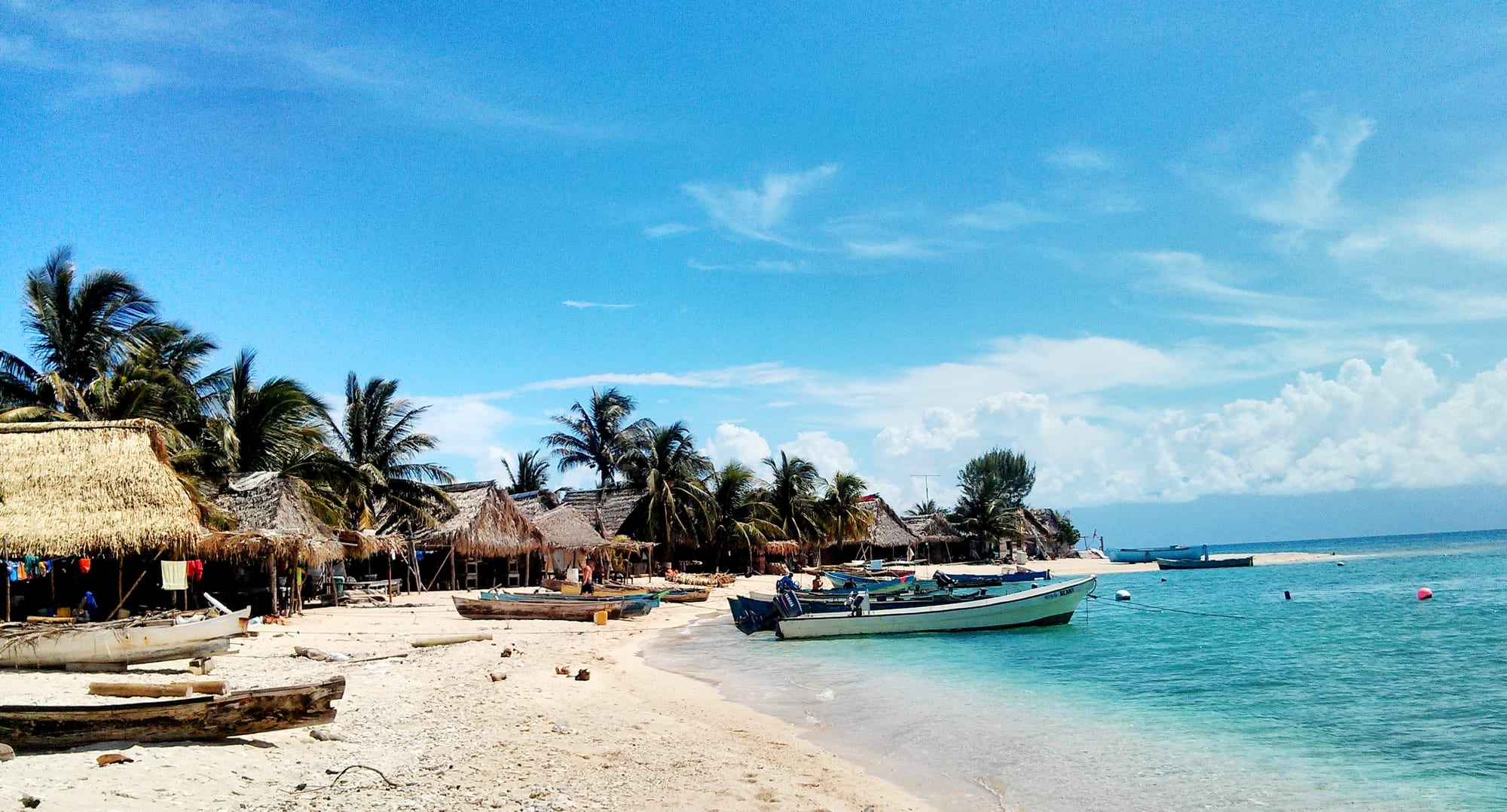 A white sand beach lined with thatched huts and palm trees, with small boats pulled up on the sand and moored in clear turquoise water under a blue sky