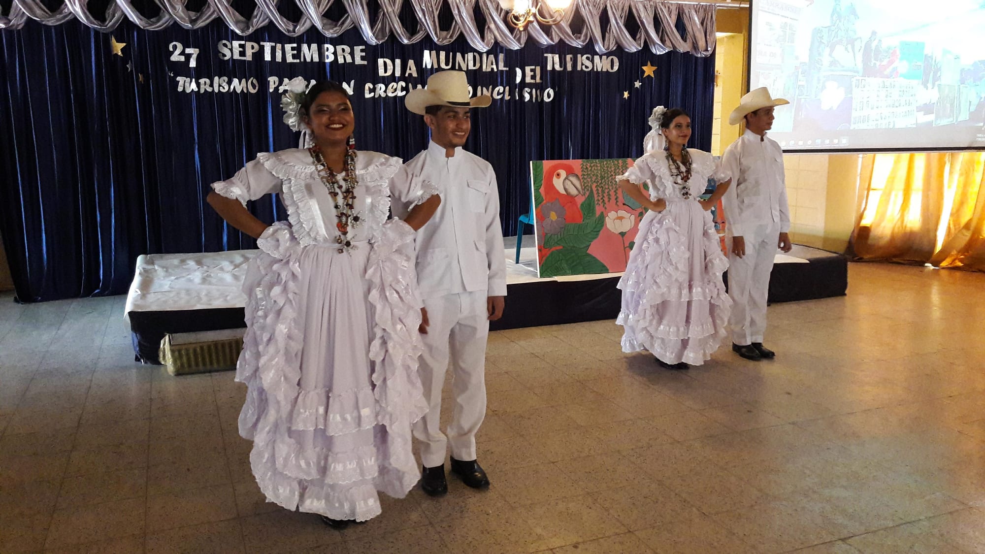 Two young women in white dresses with layers of ruffles and bead necklaces, and two young men in white shirts, trousers and cowboy hats, standing on a tiled floor in front of a stage