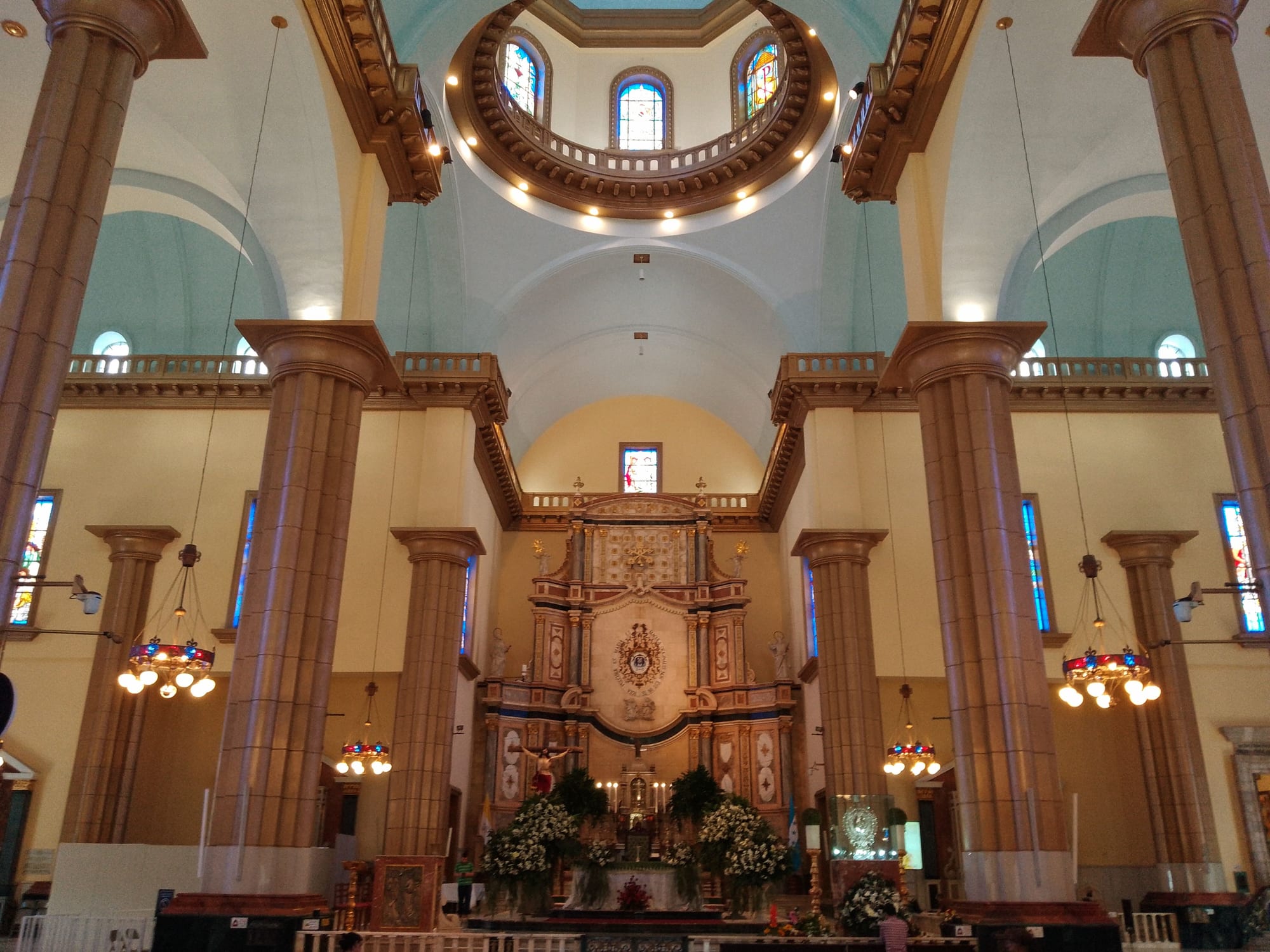 The interior of a large church with tall reddish columns, a domed ceiling ringed by stained-glass windows, and an ornate gilded altar screen banked with white flowers at the far end