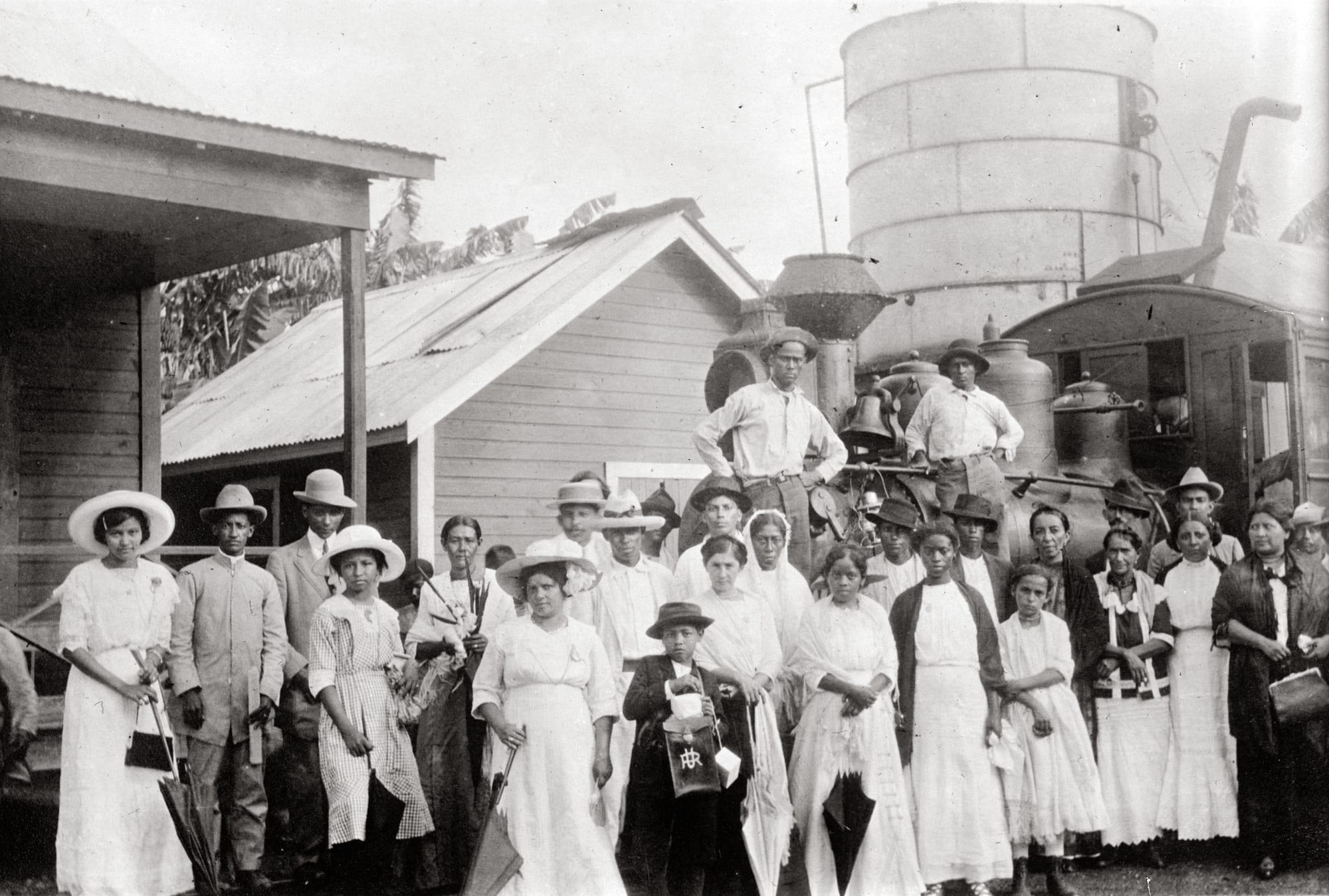 Black-and-white photograph of a crowd in white dresses, suits and straw hats posed beside a small steam locomotive, a water tank and tin-roofed wooden buildings, with banana plants behind
