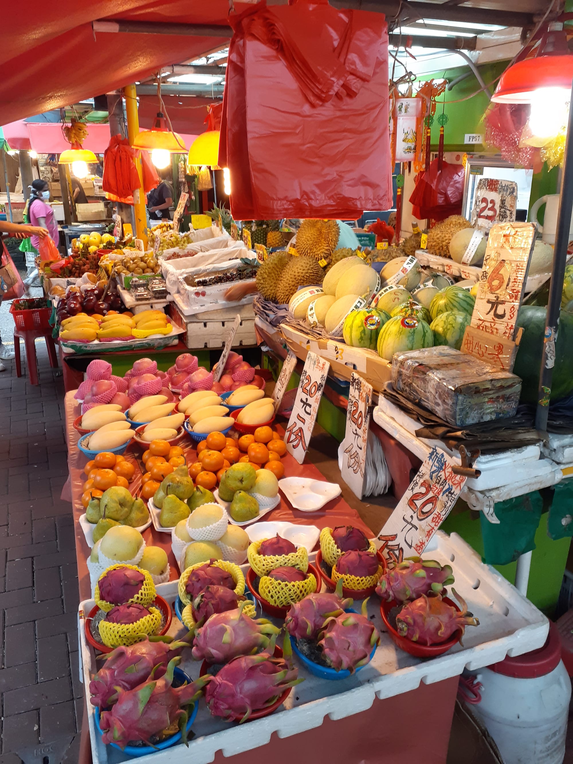 A fruit stall at an outdoor wet market in Kowloon, with baskets of mangoes, oranges, dragon fruit and melons under a red awning