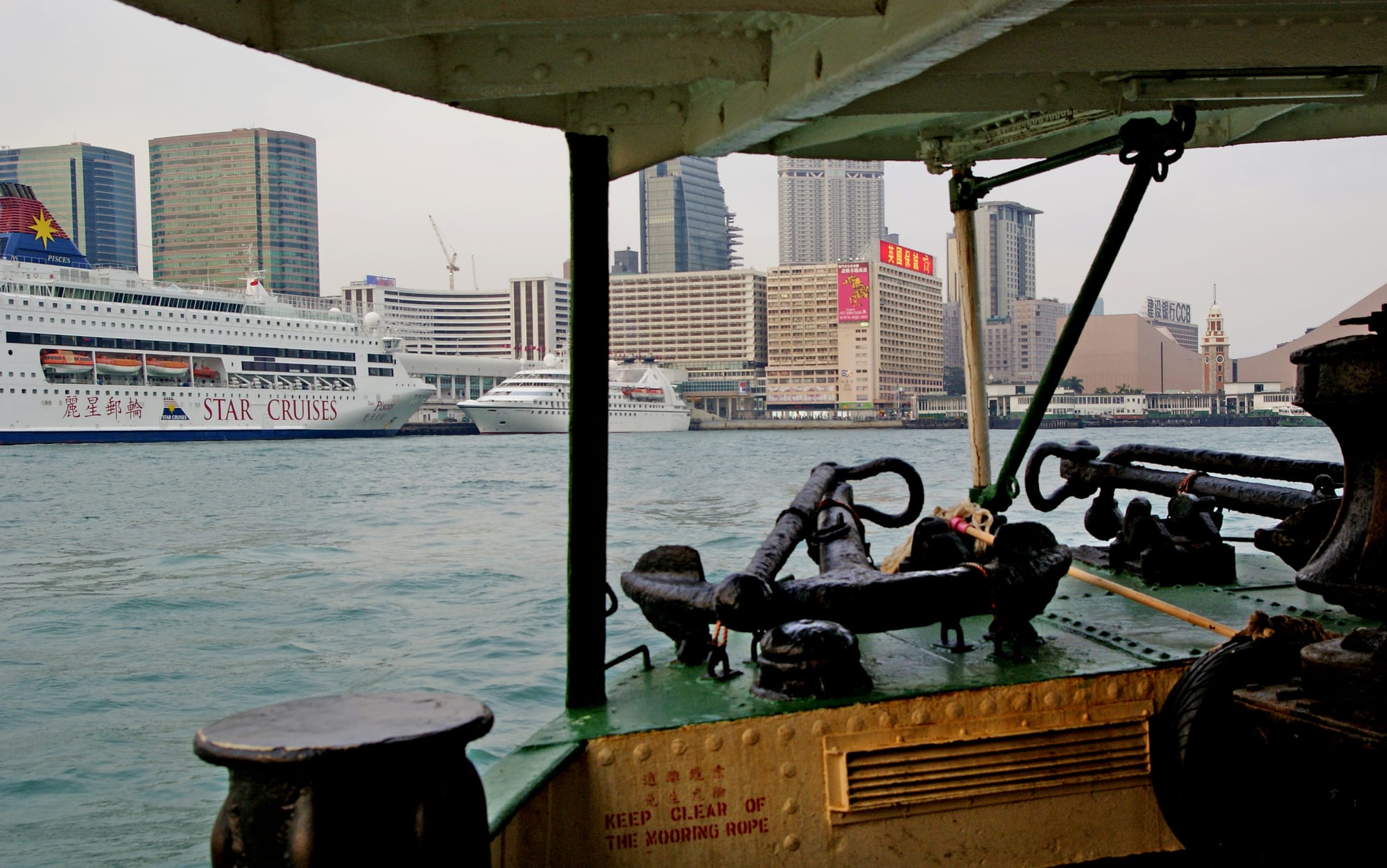 Passengers on the wooden deck of a green-and-white ferry crossing a harbour toward skyscrapers