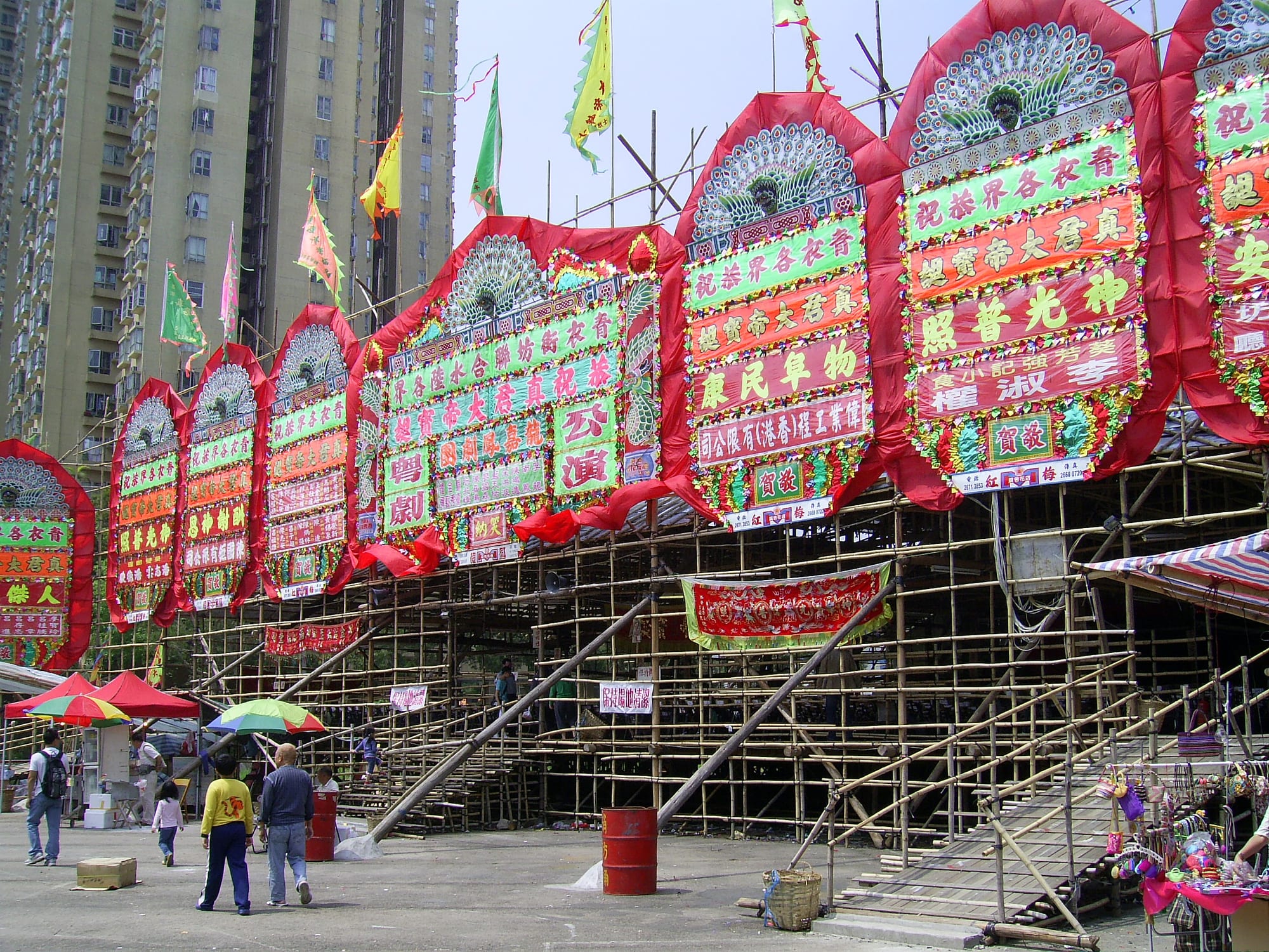 A large temporary theatre built from bamboo scaffolding, its top edge hung with tall red, green and white decorated panels covered in Chinese characters and peacock motifs