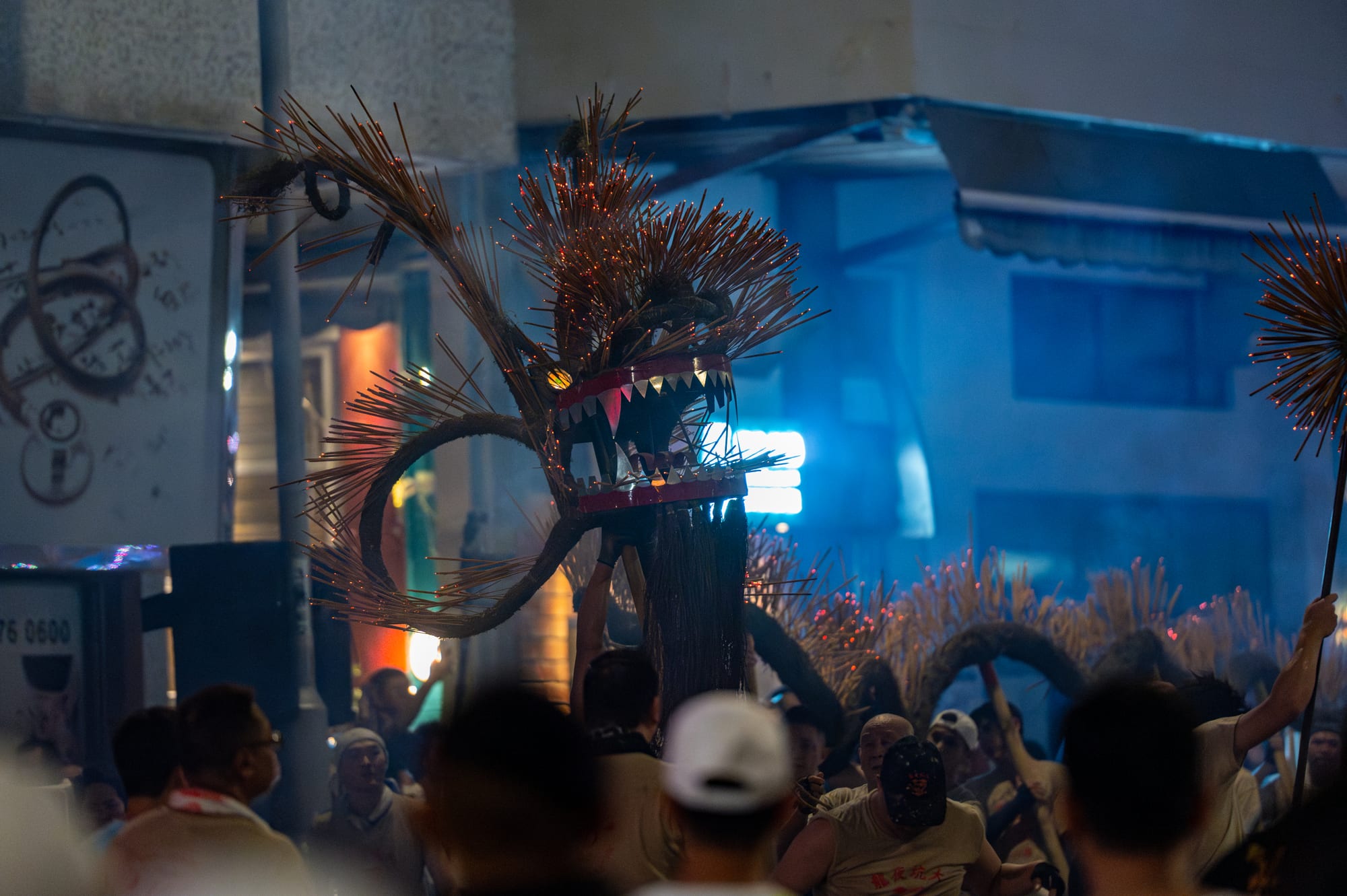 The head of a large dragon made of straw and bristling with glowing incense sticks is held aloft above a crowd on a street at night