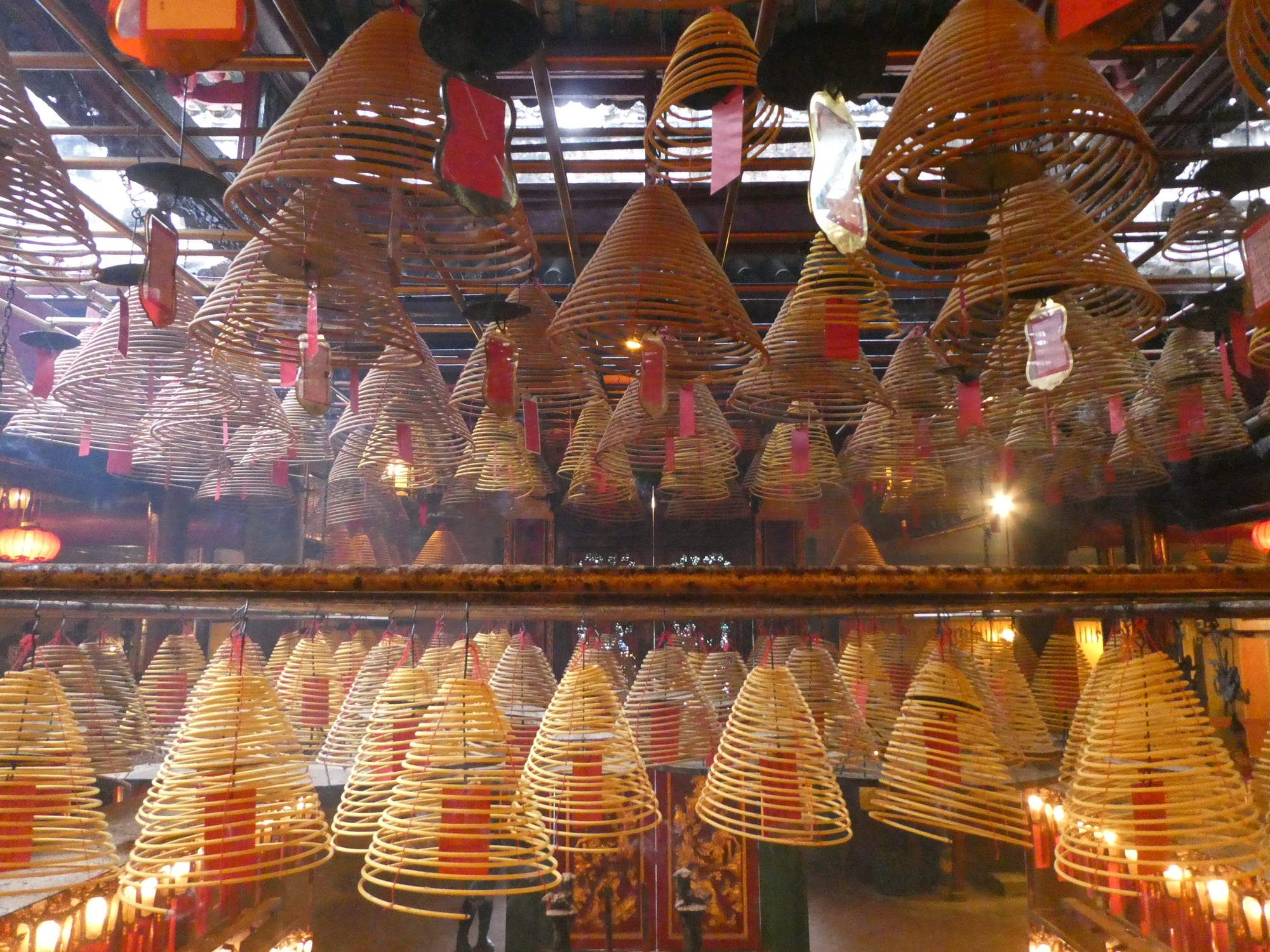 Dozens of large cone-shaped incense coils hang in tiers from the ceiling of a temple, each with a red paper tag, glowing in the dim light